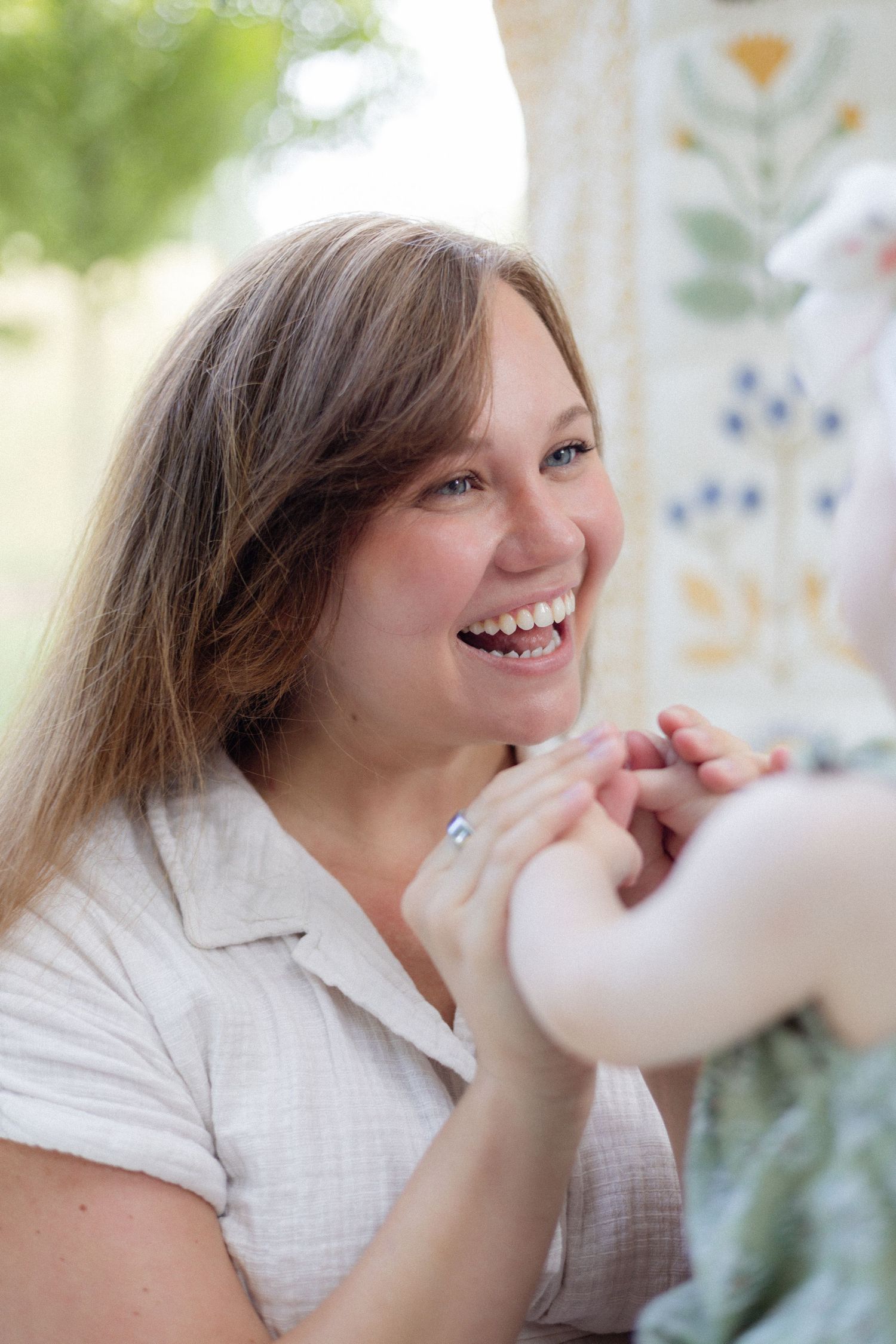 Smiling woman with long brown hair laughing joyfully while holding a baby's hands near a floral backdrop.