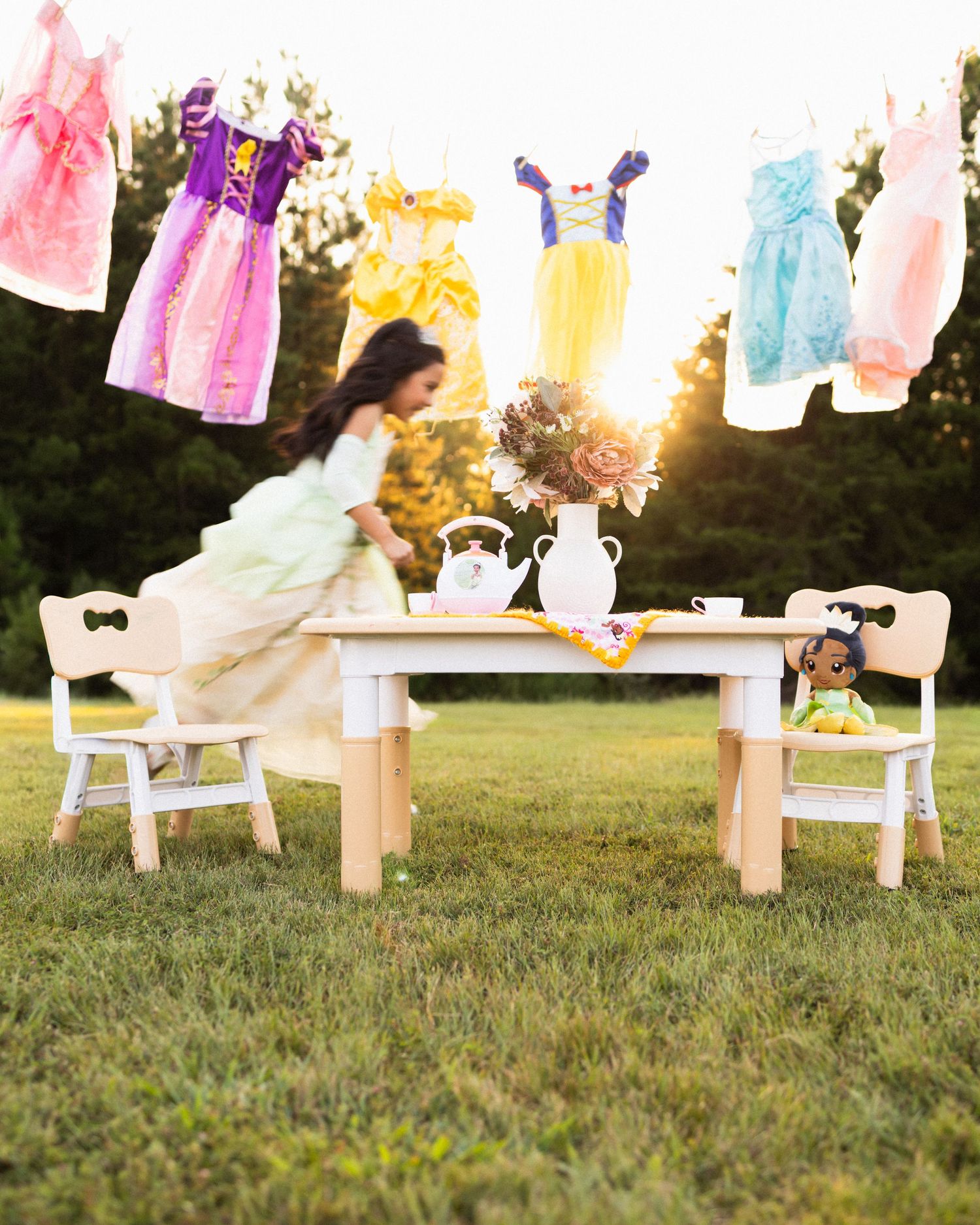 Girl in yellow princess dress at outdoor tea party table with princess dresses hanging on clothesline at sunset.
