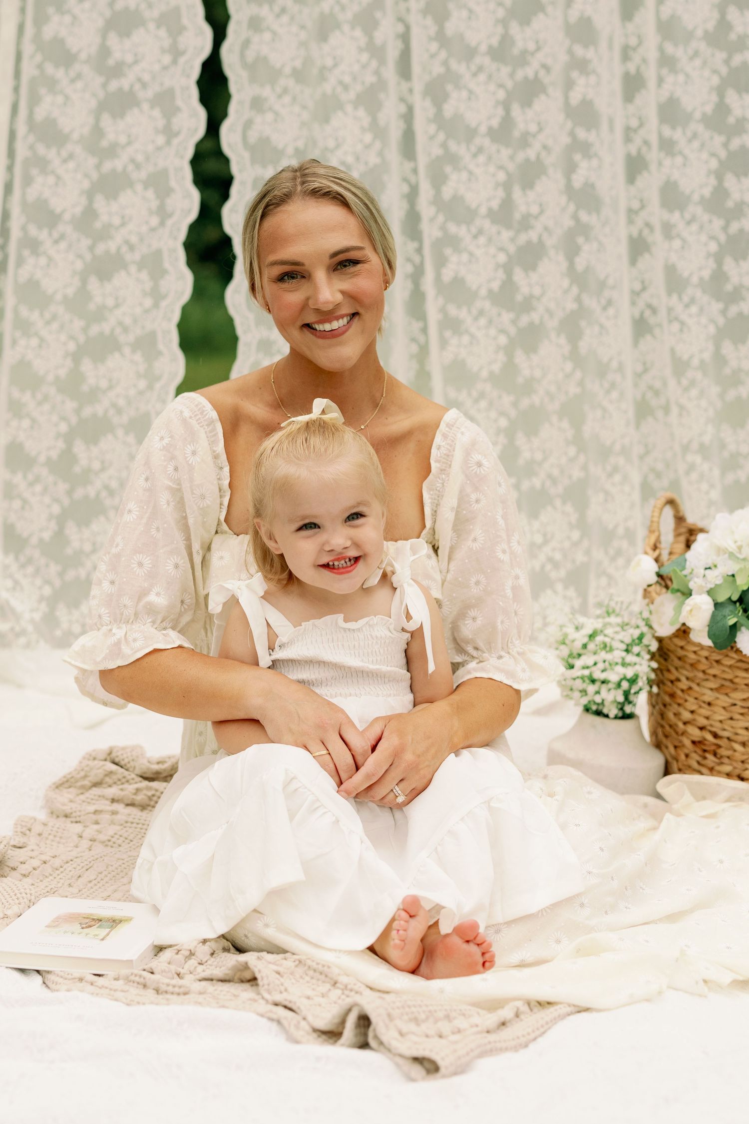 Smiling blonde mother in white lace dress sitting with happy toddler girl on cozy blanket indoors.
