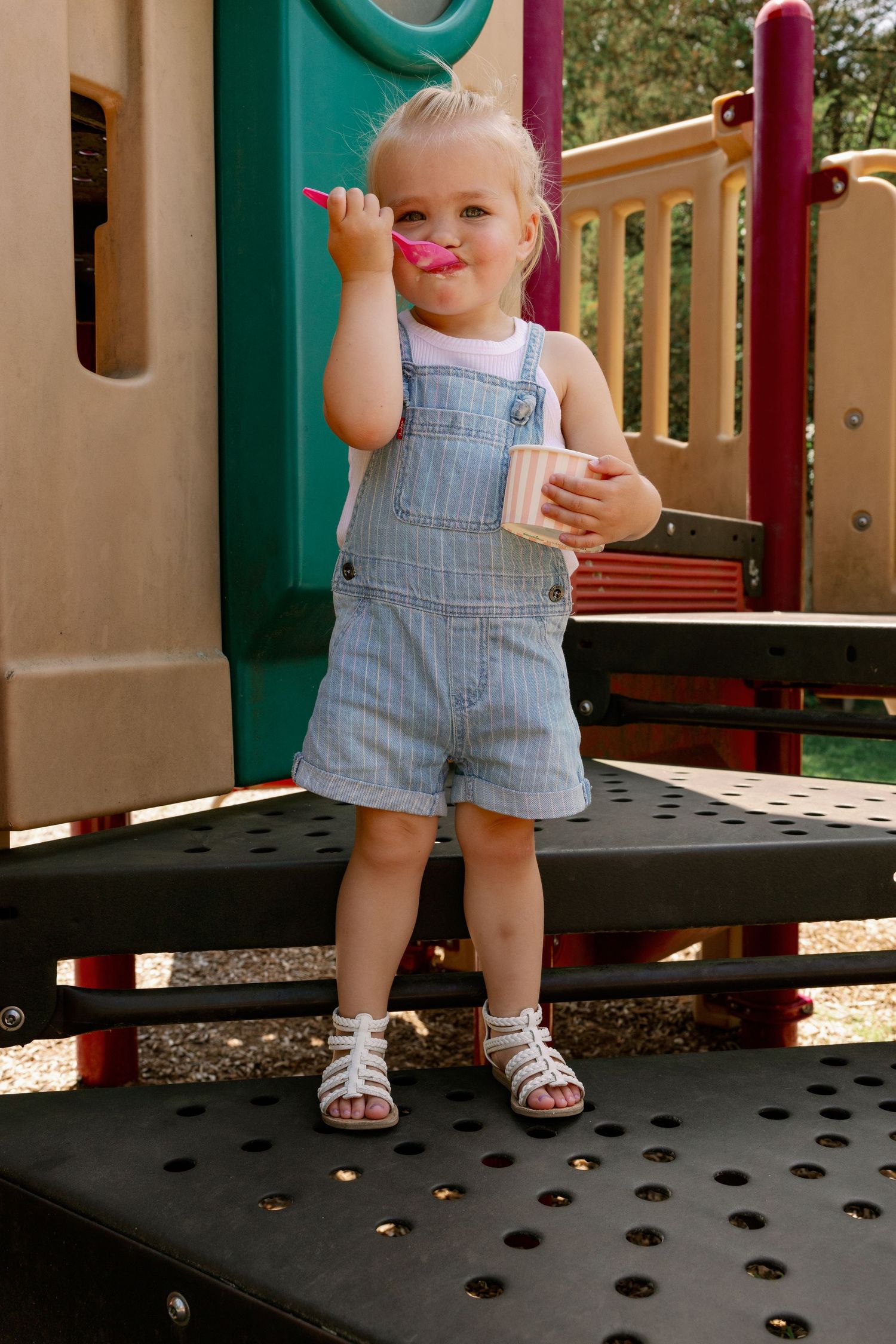 Adorable toddler in denim overalls enjoying ice cream on a playground structure.
