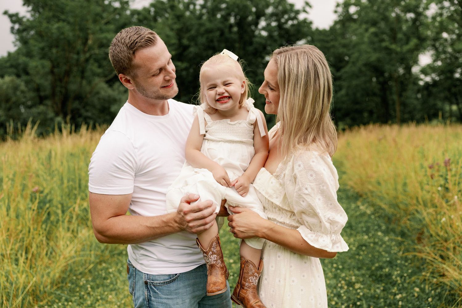 Happy family with parents holding their smiling toddler daughter in a sunny green meadow.