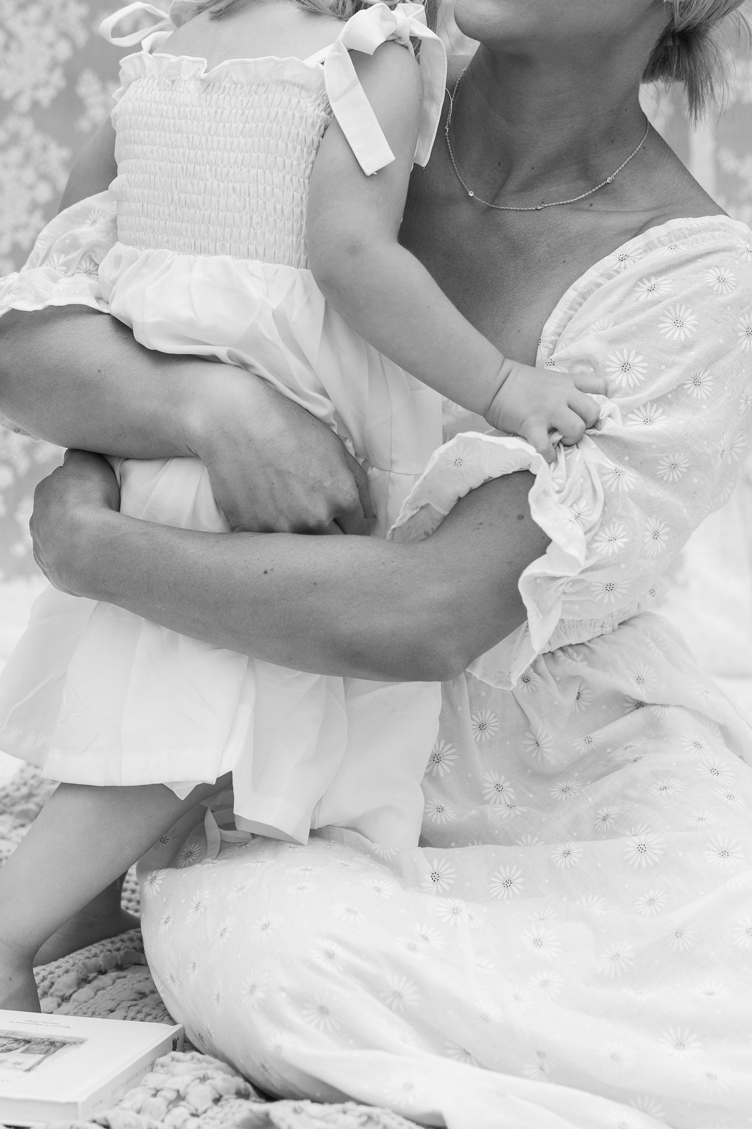 Black and white close-up of mother holding toddler girl in white smocked dress with bow detail.