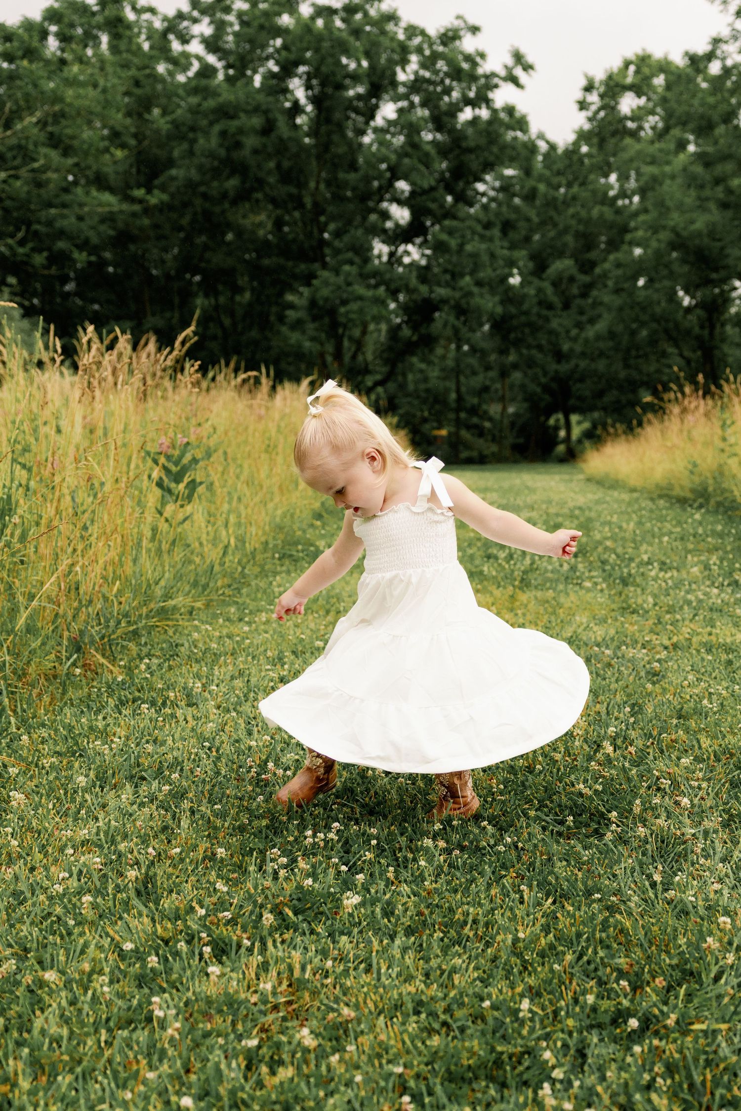 Adorable toddler girl in white dress and cowboy boots twirls in a green meadow.