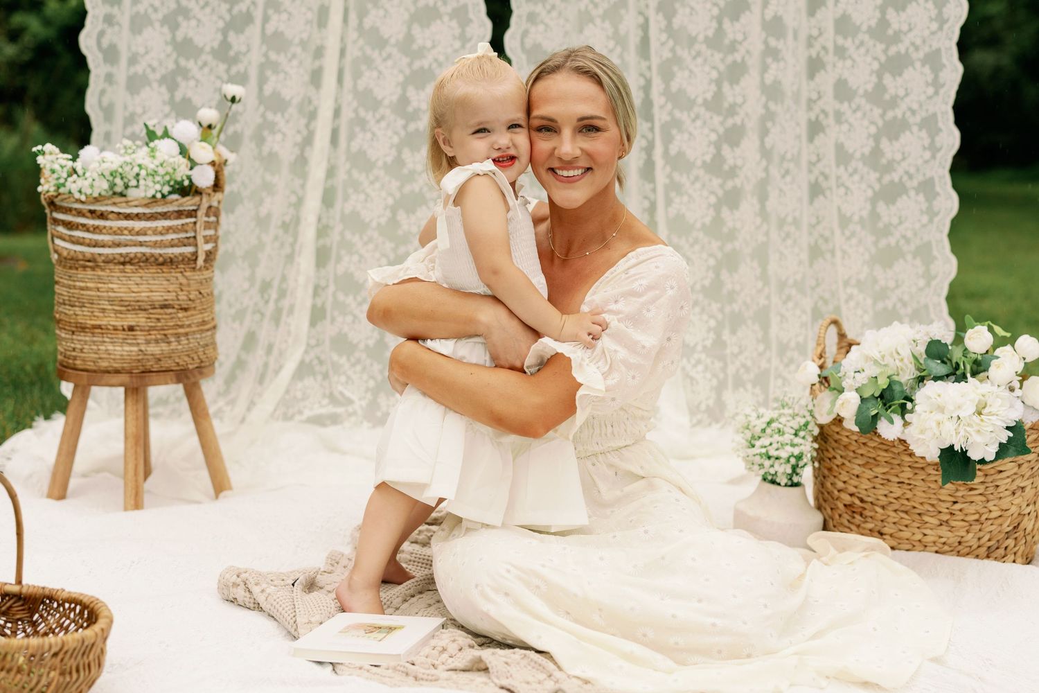 Mother in white dress hugging toddler girl outdoors on white blanket with lace backdrop and wicker baskets.