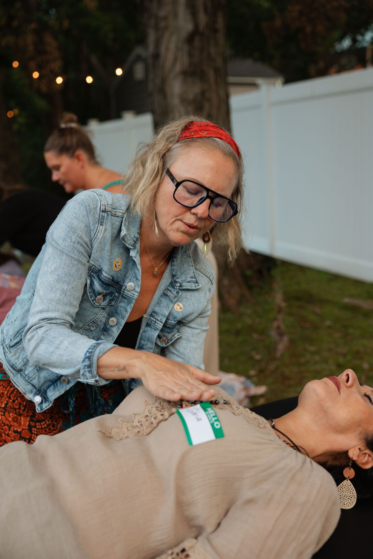 Woman with glasses and red headband performing energy healing on a person lying down at an outdoor evening event.