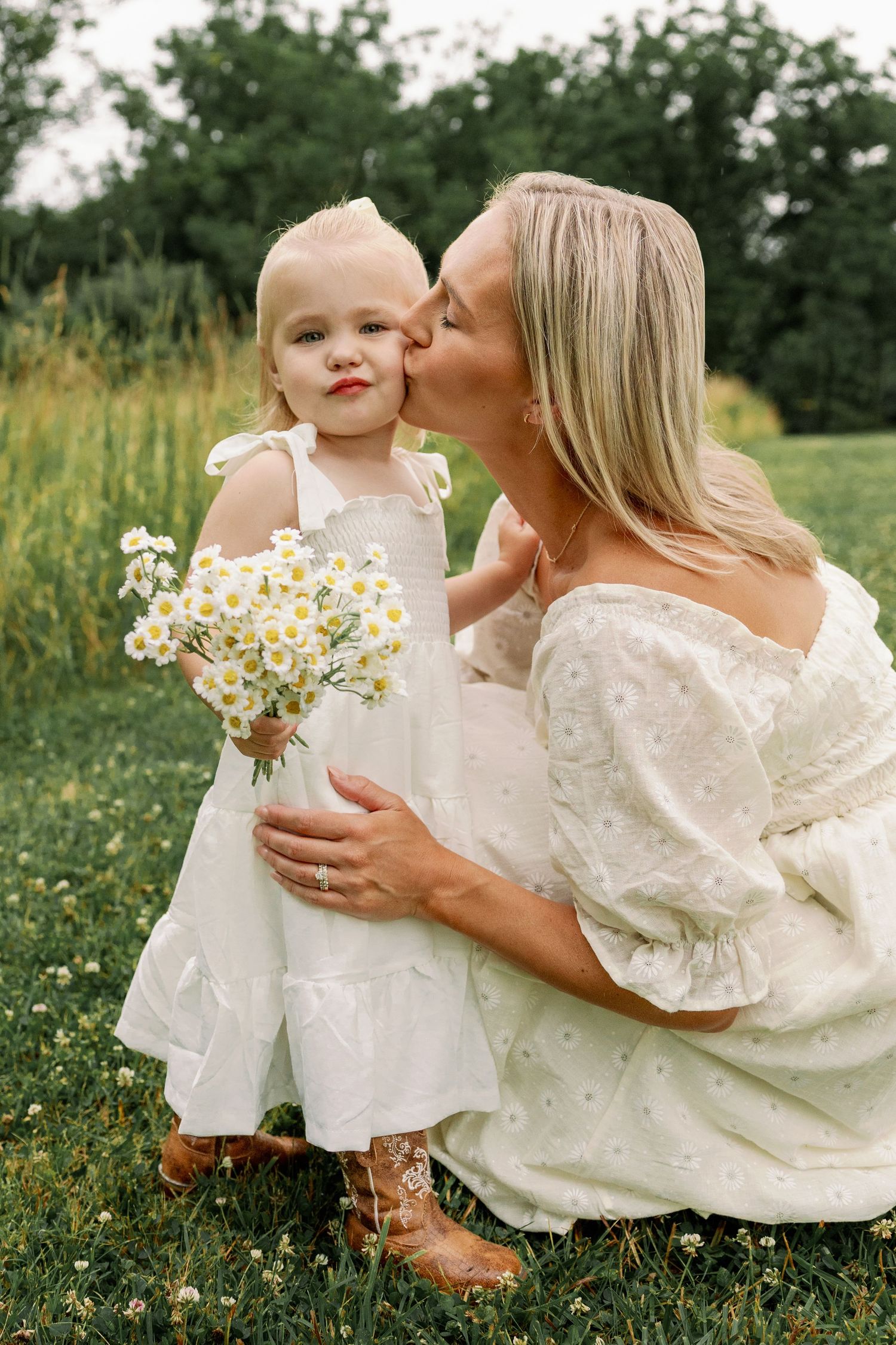 Blonde mother kisses her toddler daughter holding a bouquet of white wildflowers outdoors.