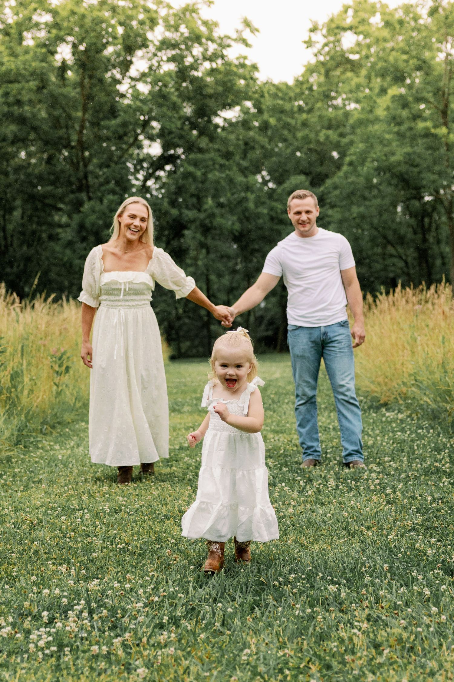 Joyful family of three holding hands with excited toddler in white dress in a clover field.