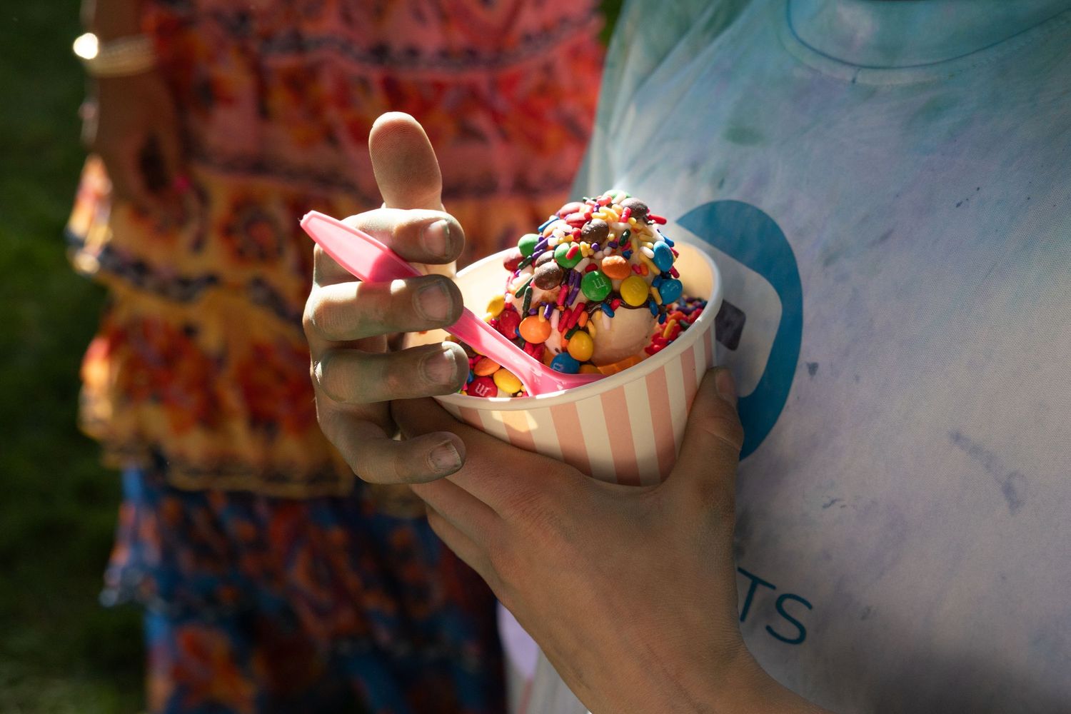 Person holding a colorful sprinkle-topped ice cream cup at an outdoor summer event.