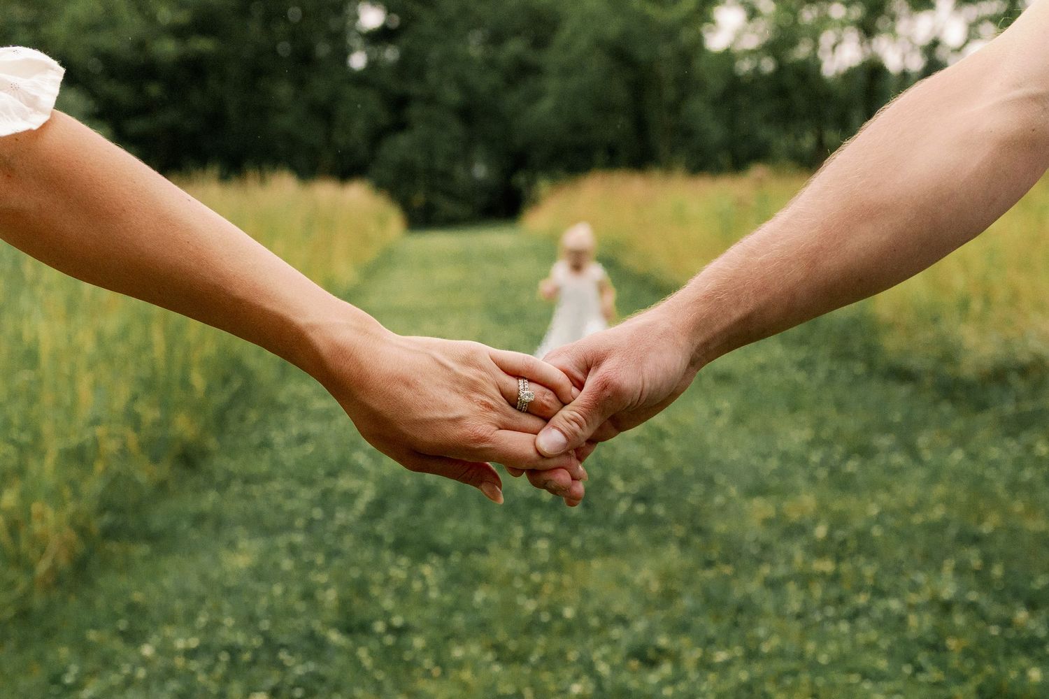 Close-up of couple holding hands in a green field with a child in a white dress visible in the background.