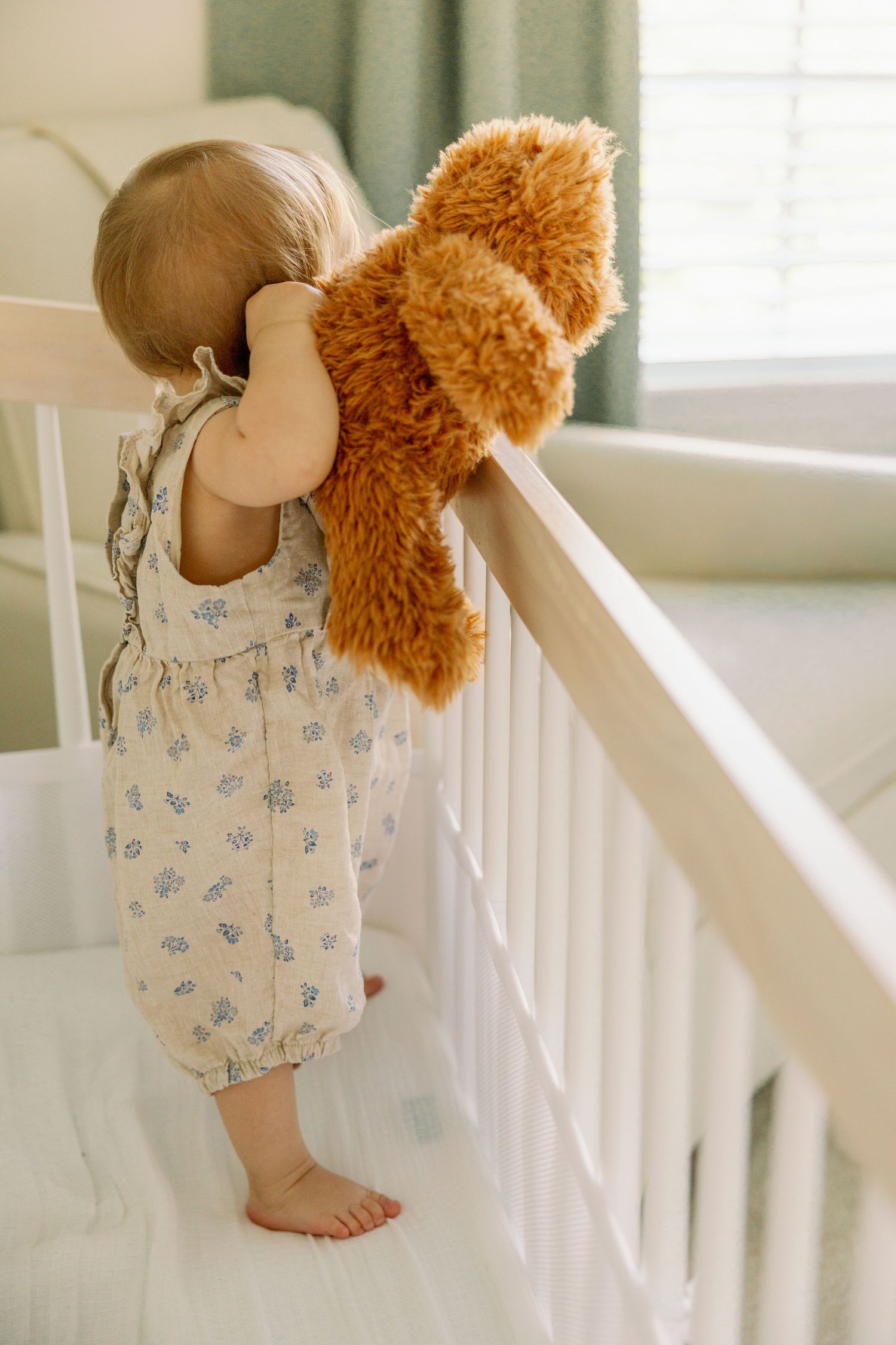 Baby standing in white crib holding a brown teddy bear near a sunny window.