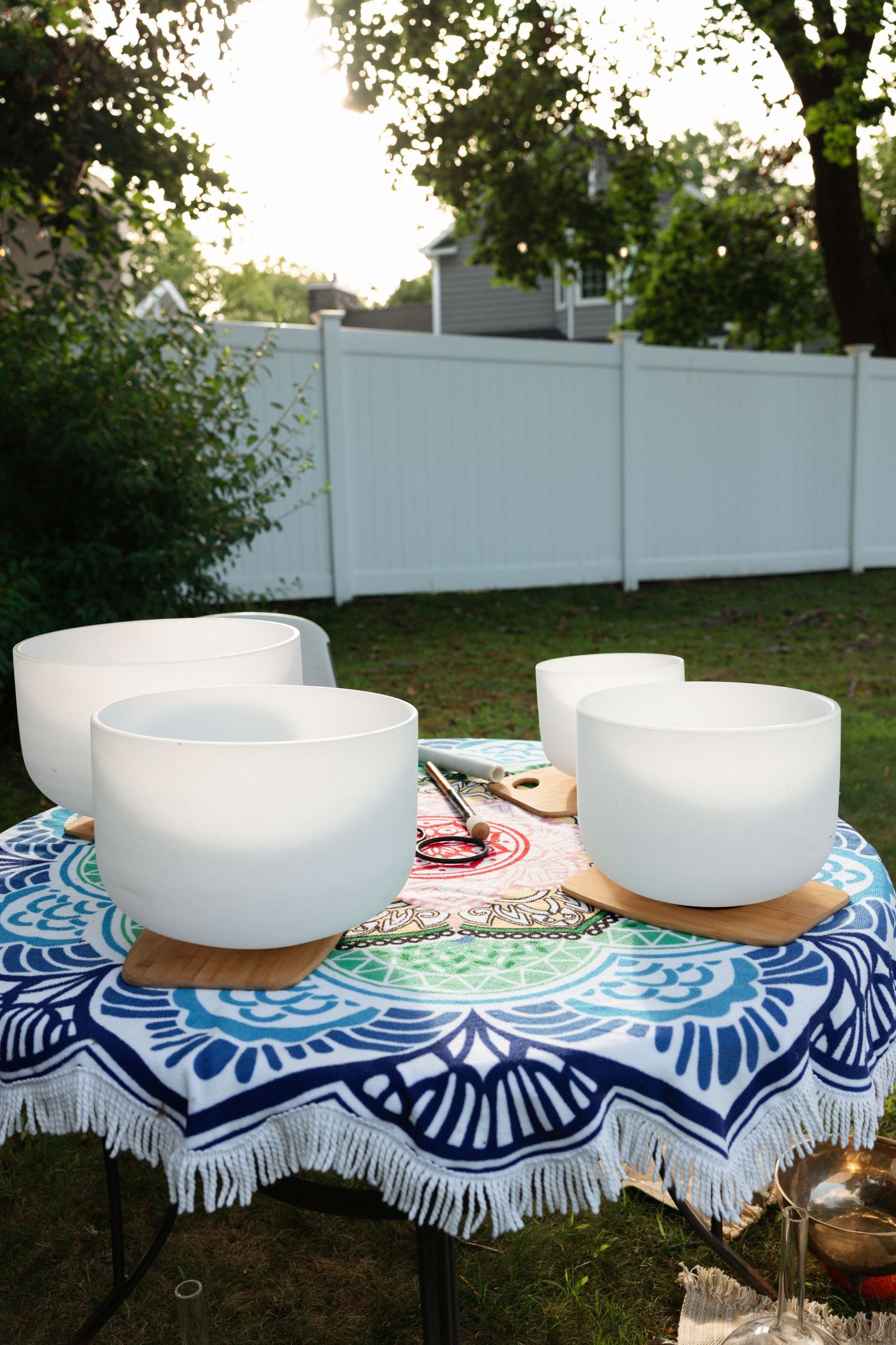 Four white crystal singing bowls arranged on a colorful mandala blanket on a table in a serene backyard setting.