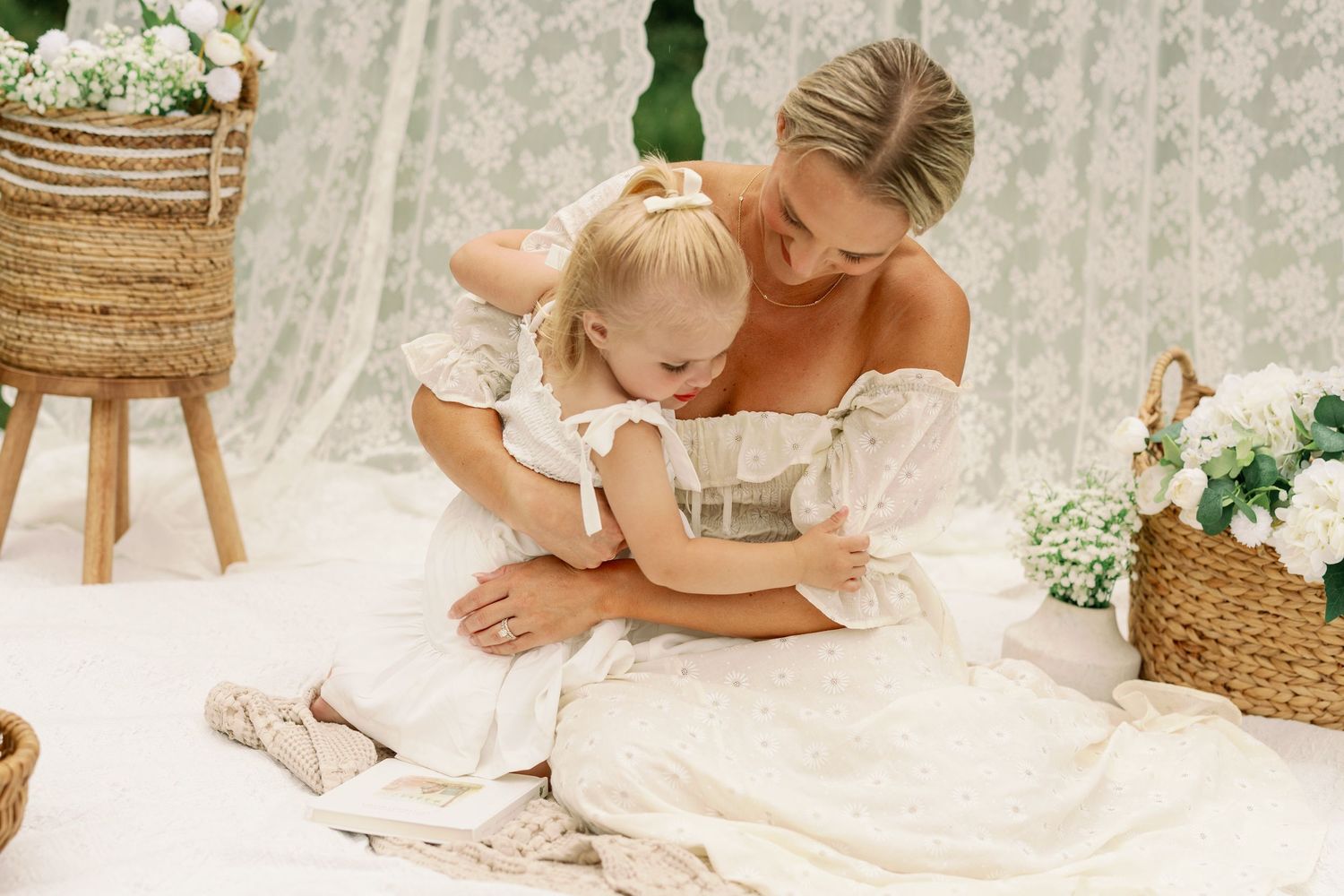 Mother and young daughter in matching white dresses sitting together on a blanket in a boho-styled setting.