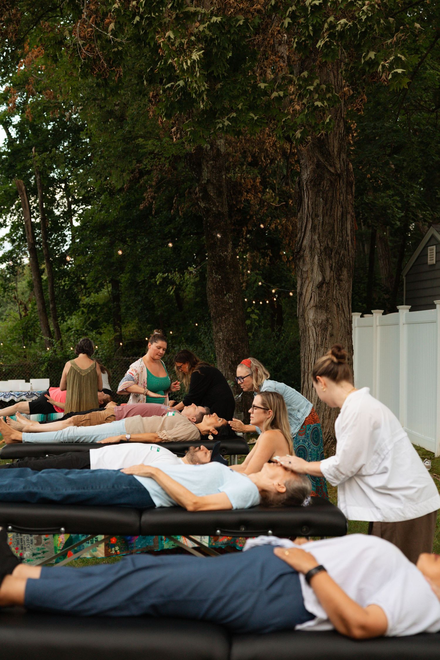 Outdoor group massage session with people lying on tables while therapists work on them, surrounded by trees and string lights.