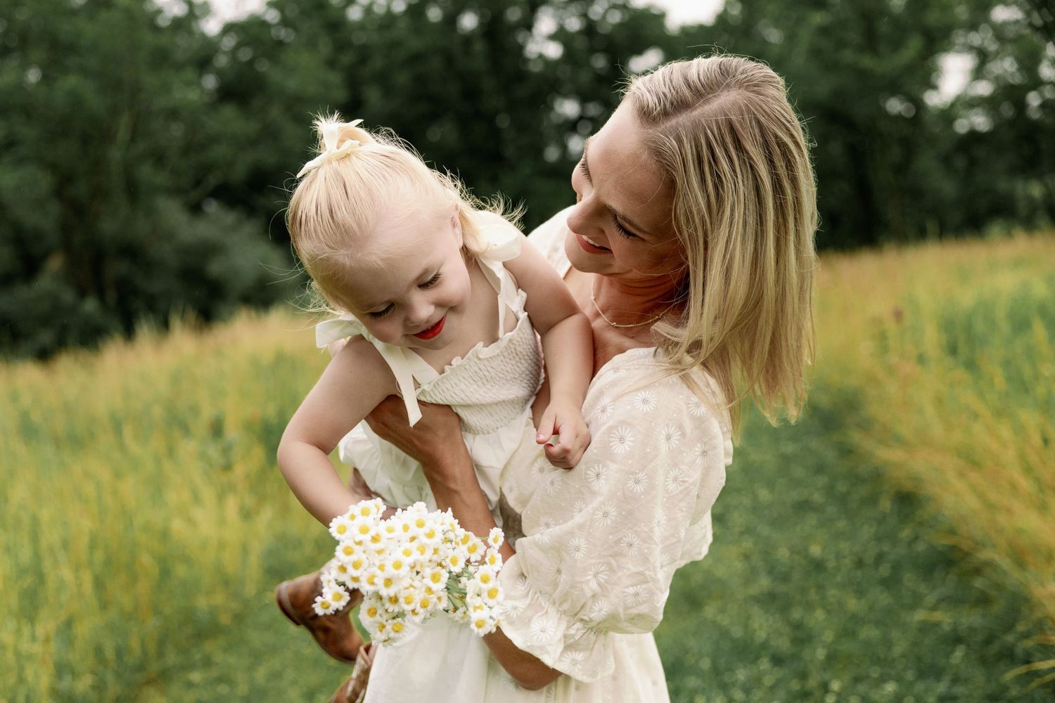 Joyful mother in white dress lifting and hugging her laughing toddler daughter holding white daisy flowers.