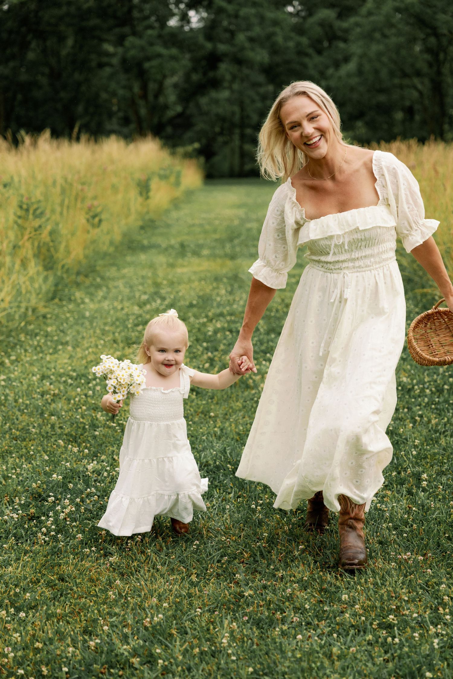 Laughing mother in white dress holding hands with her smiling toddler daughter in a wildflower meadow.