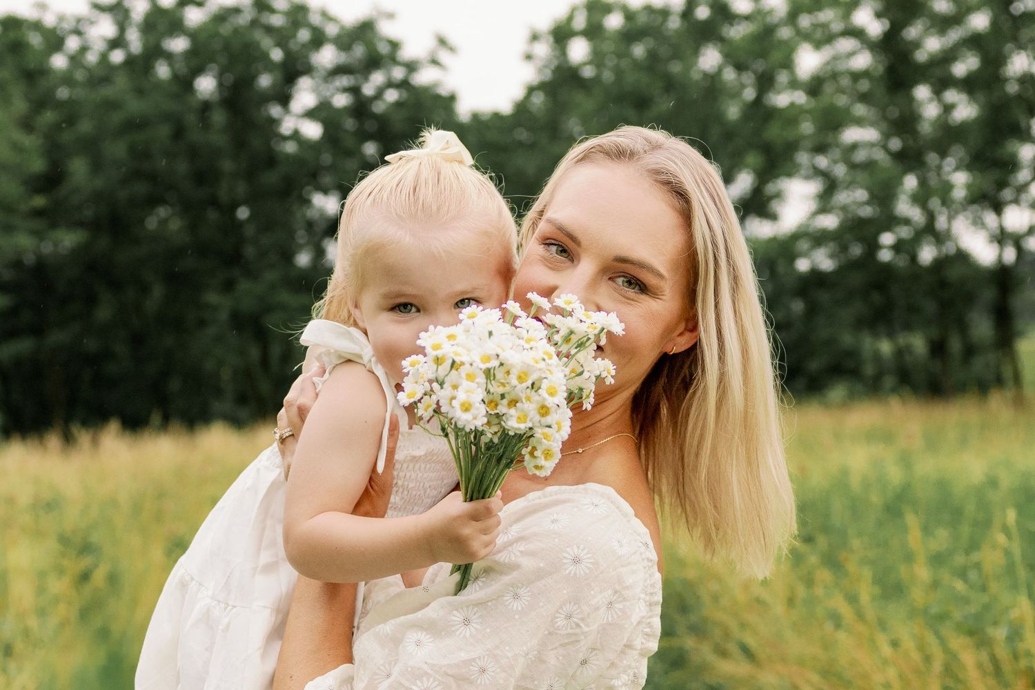 Blonde mother holding young daughter in white dress as child presents a bouquet of white daisies outdoors.
