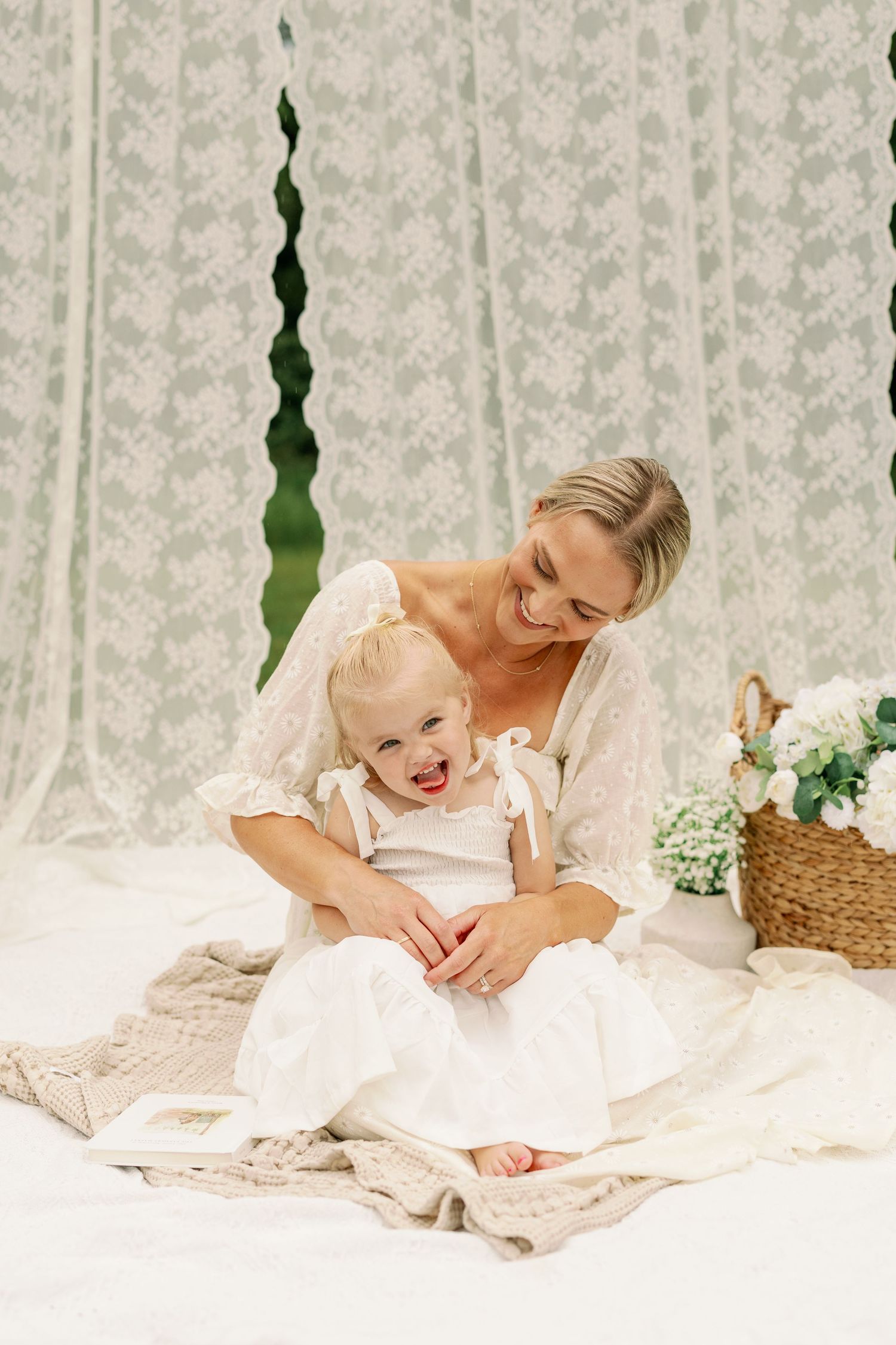 Laughing blonde mother cuddles giggling toddler girl in white dress on cozy blanket with lace backdrop and flowers.