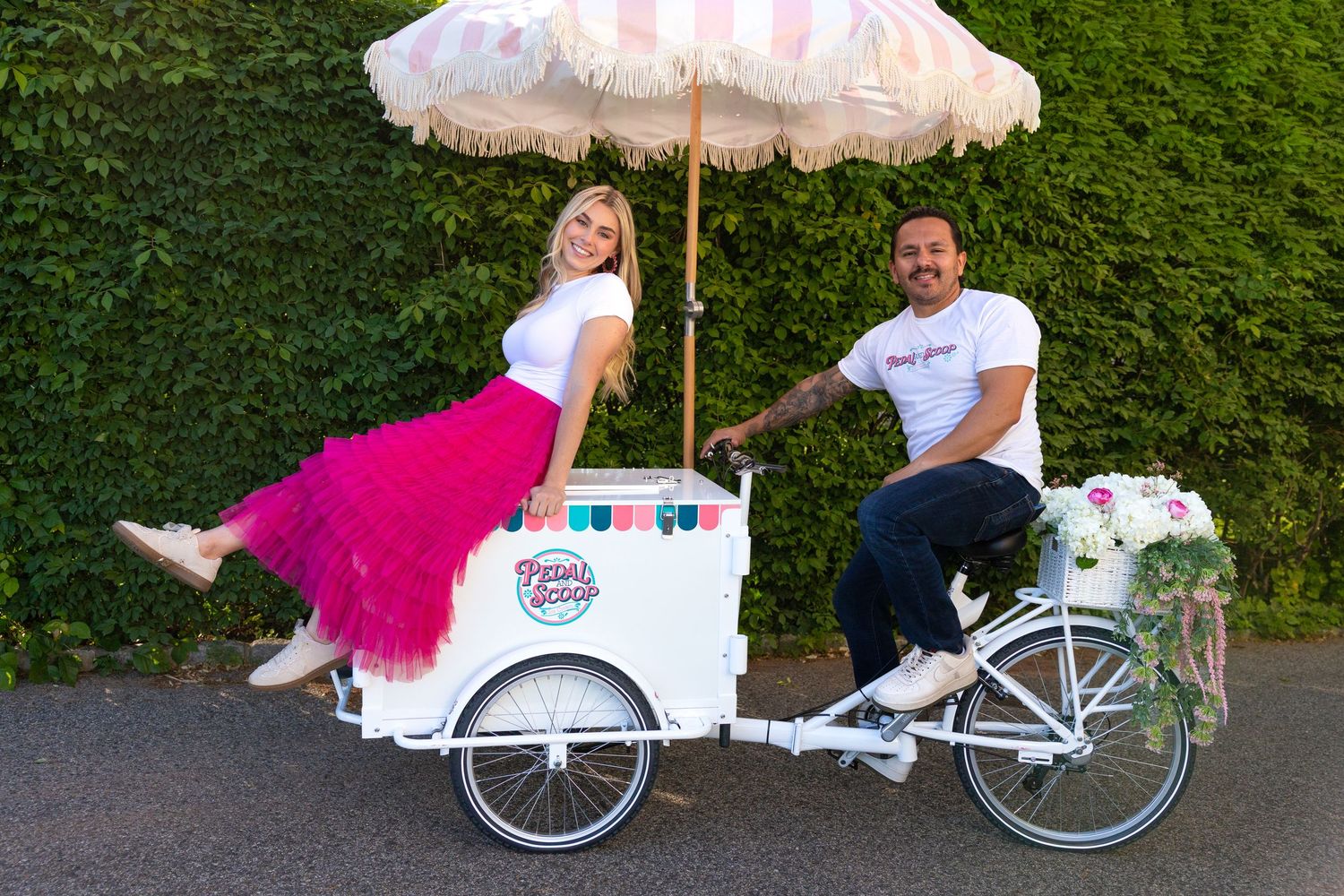 Man and woman posing with a white vintage ice cream tricycle cart under a striped umbrella outdoors.