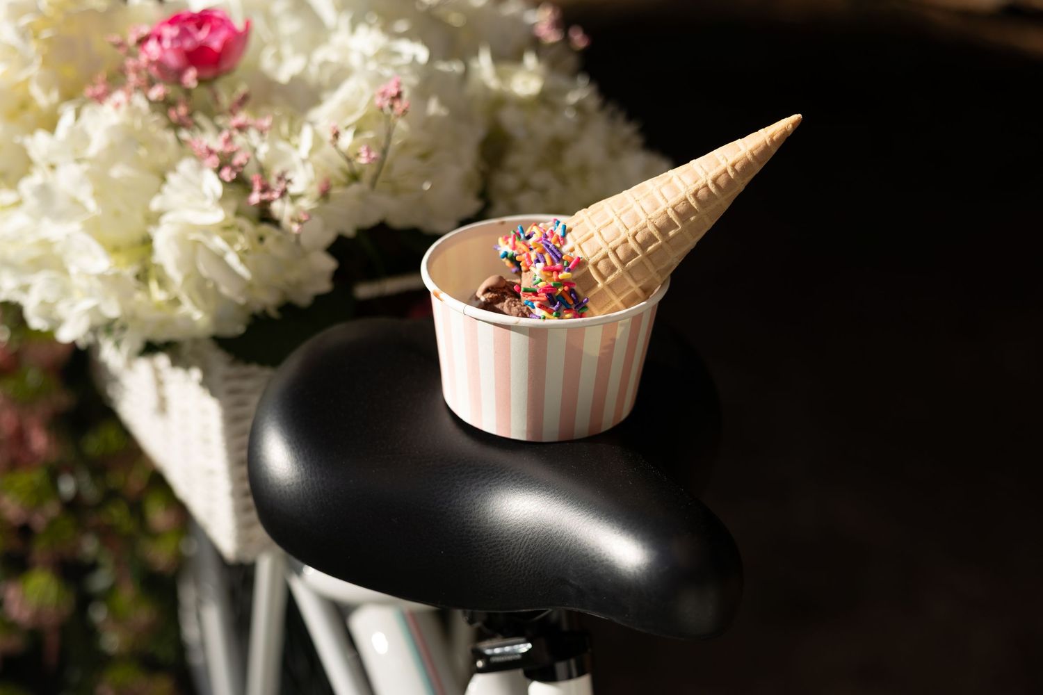 Ice cream cone with rainbow sprinkles resting in a striped cup on a black bicycle seat outdoors.