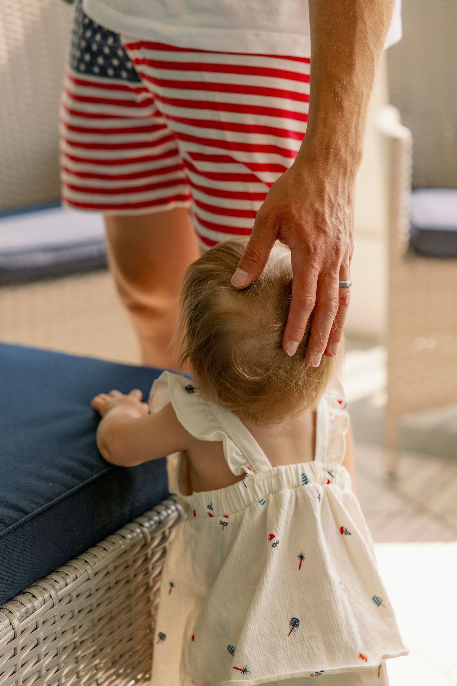 Father in American flag shorts gently touching toddler daughters head as she stands by outdoor furniture.