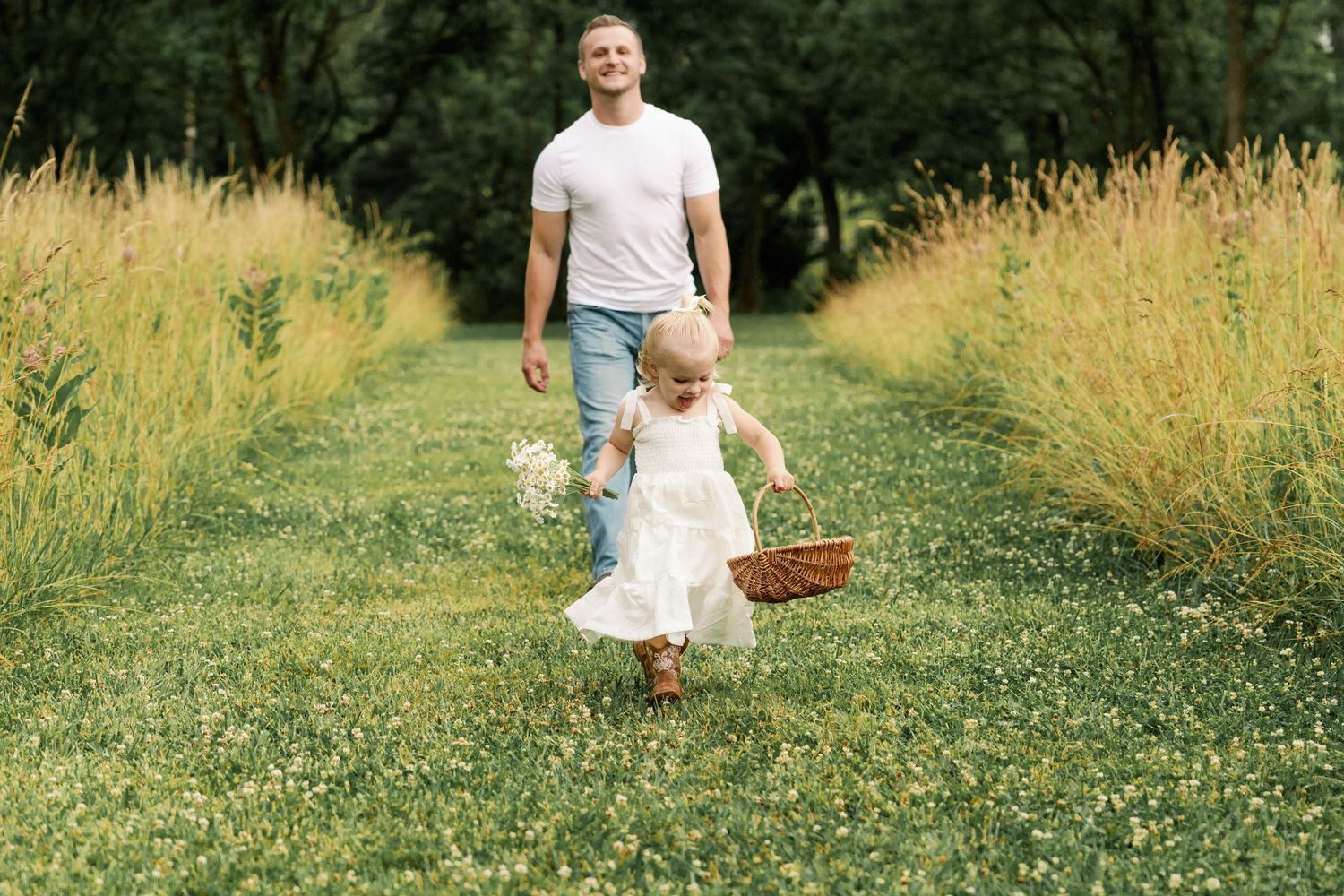 Father in white t-shirt and jeans walks behind toddler daughter carrying basket in green meadow.