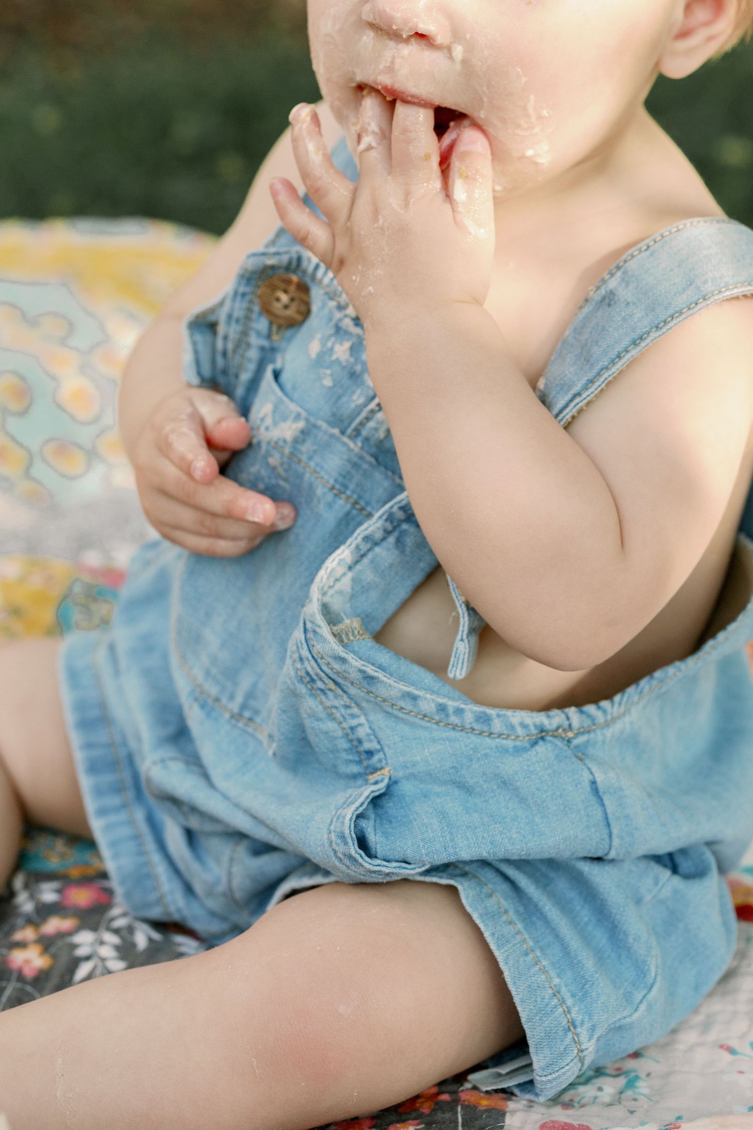 Close-up of baby in denim overalls eating cake with frosting on hands and face.