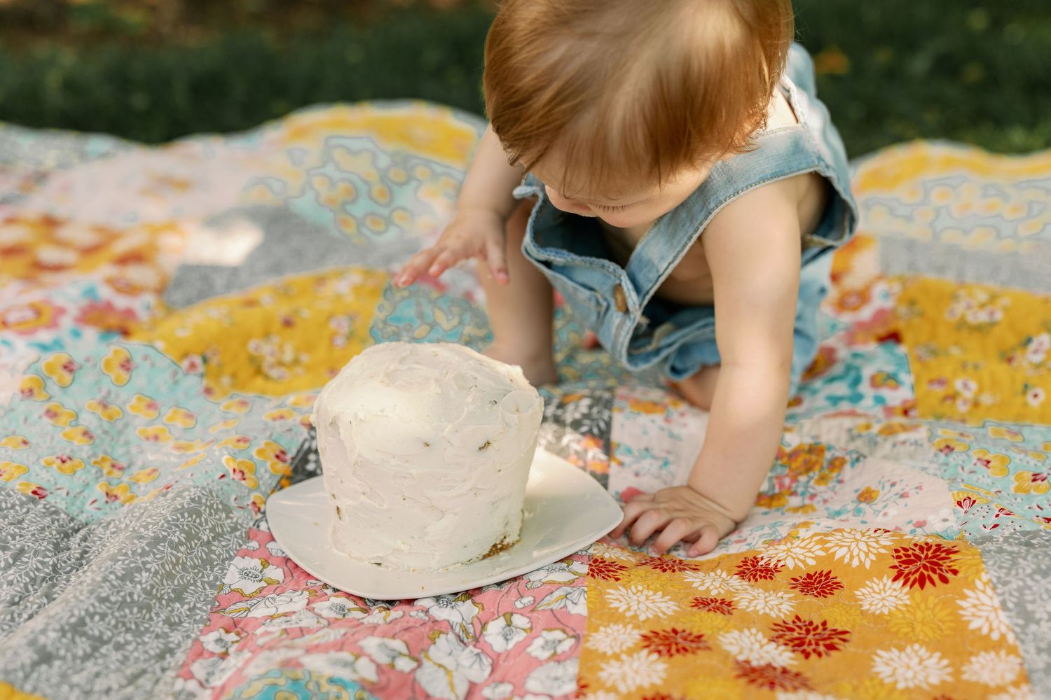 Toddler in denim overalls reaching toward a smash cake on a colorful patchwork quilt outdoors.