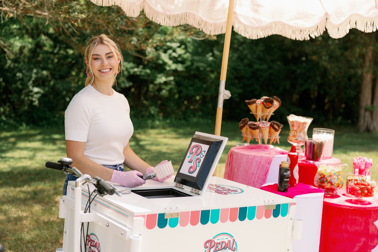 Young woman working at a colorful Pedal Scoop ice cream cart in a lush garden setting.