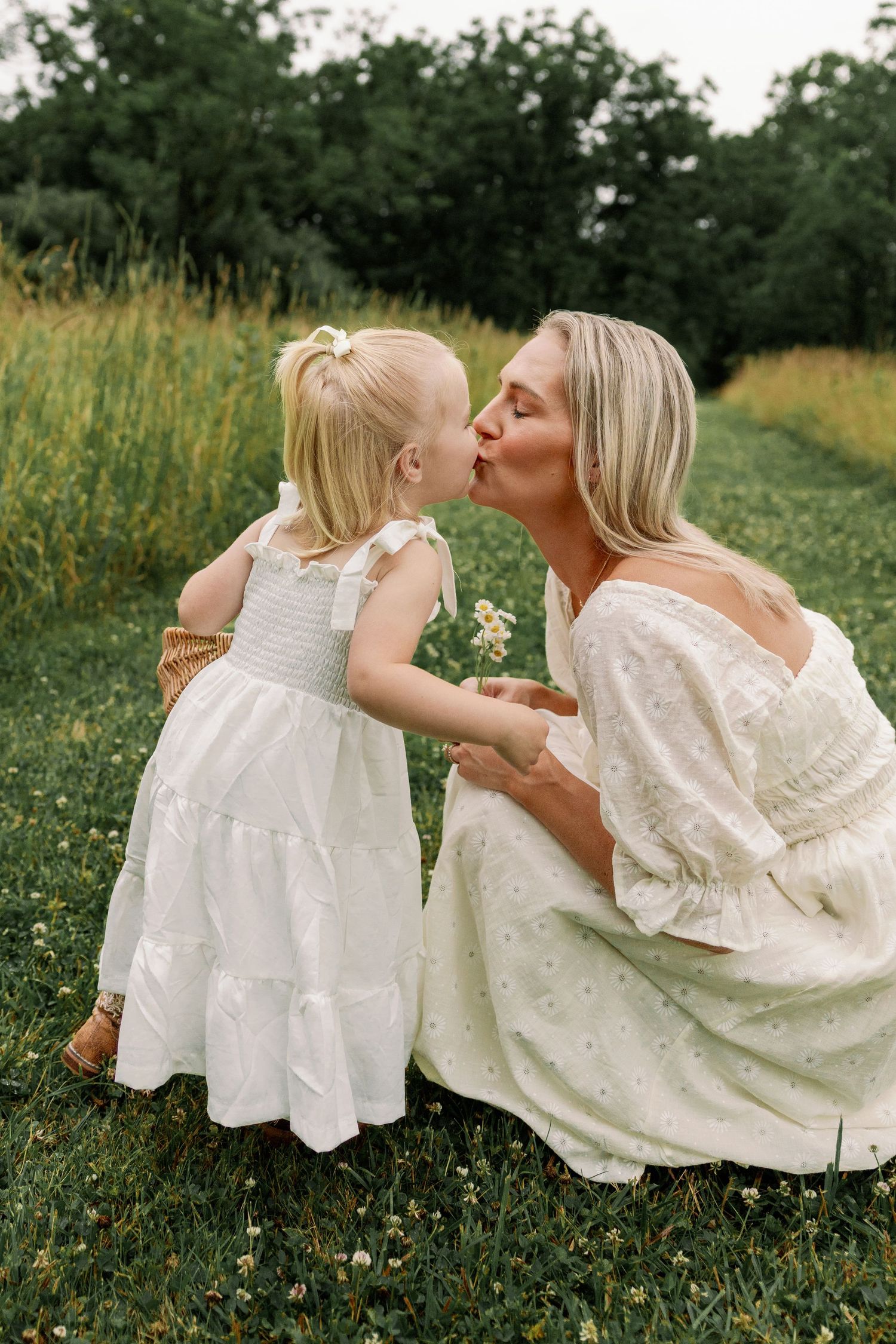 Mother and toddler daughter in matching white dresses share a sweet kiss in a lush green meadow.