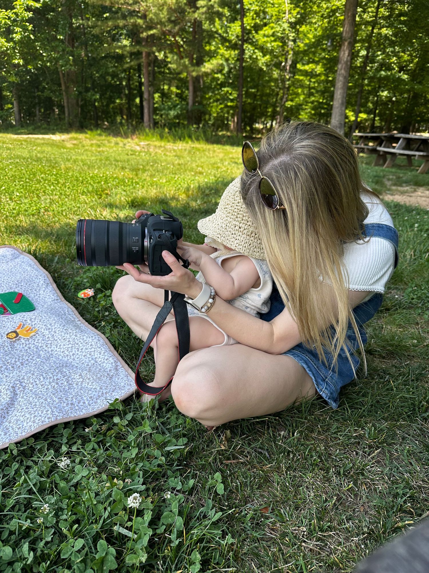 A young blonde woman sits on grass photographing with a DSLR camera outdoors in a sunny park setting.