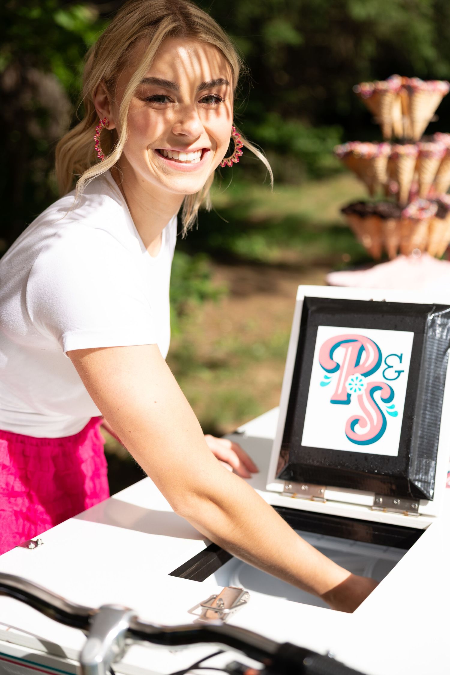 Smiling woman leaning over a white ice cream cart displaying a decorative pink and blue logo sign.