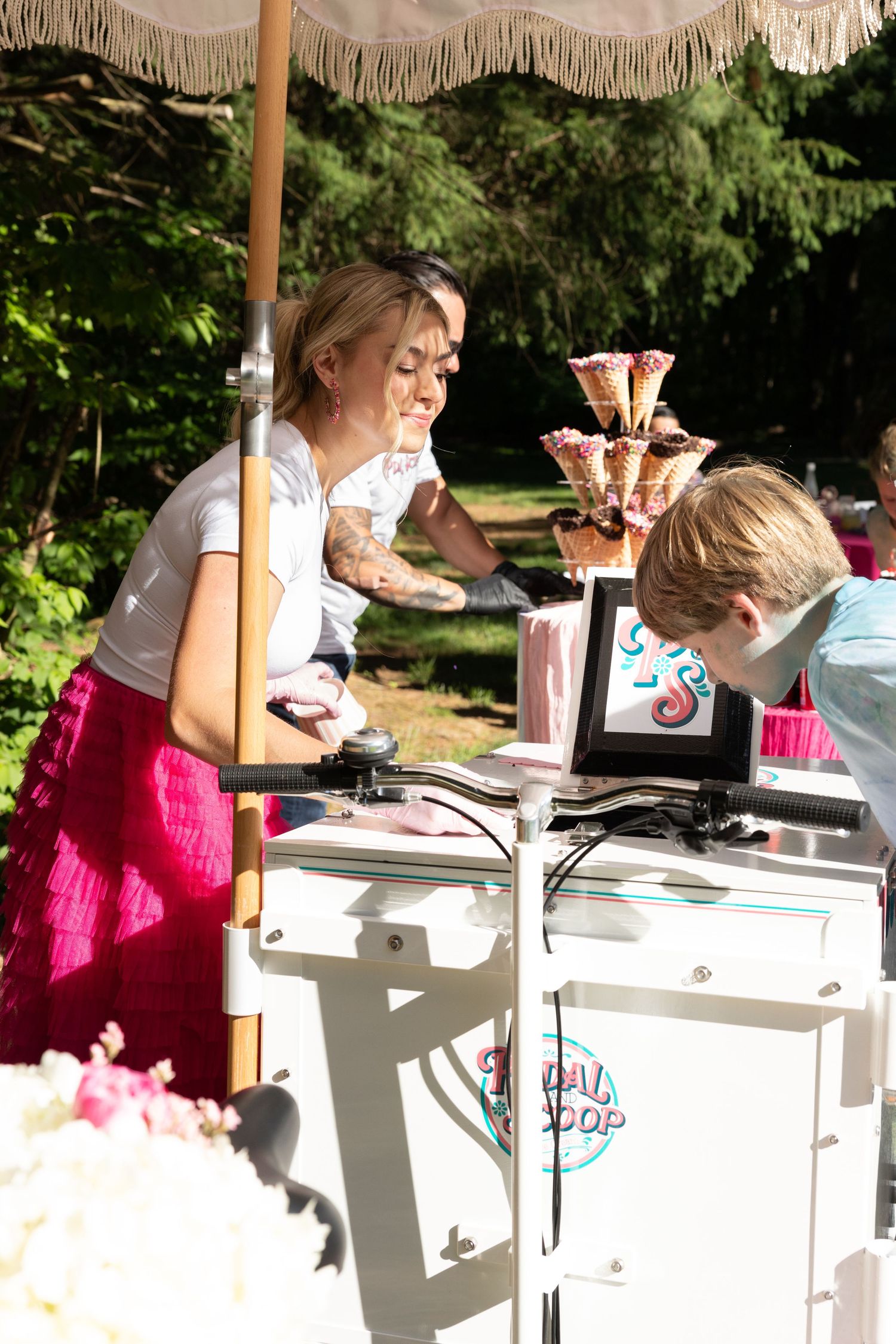 Smiling woman in pink skirt serving ice cream to a child at an outdoor dessert cart.