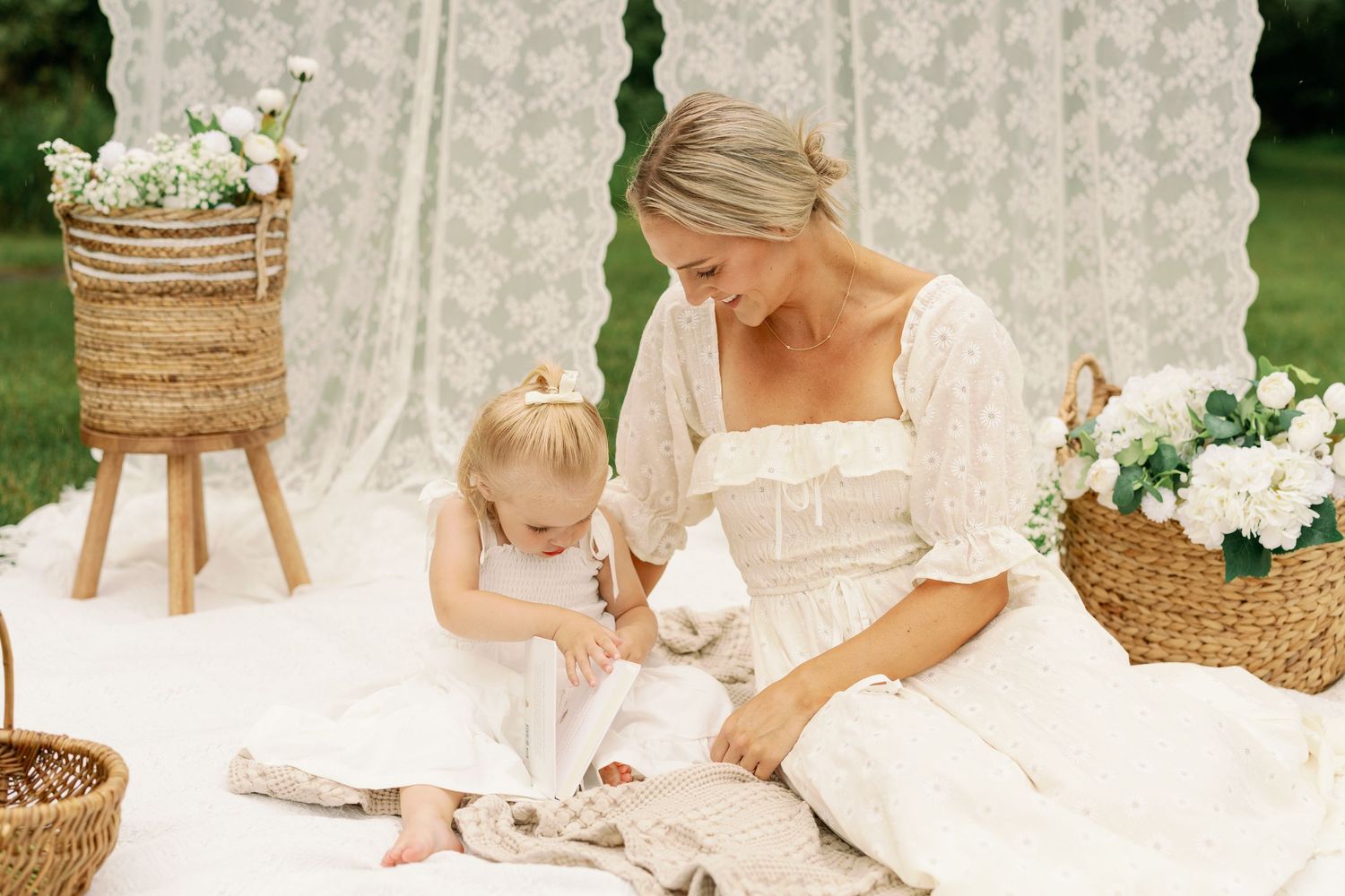 Mother in white dress reading with toddler daughter on white blanket with lace backdrop and floral decor.