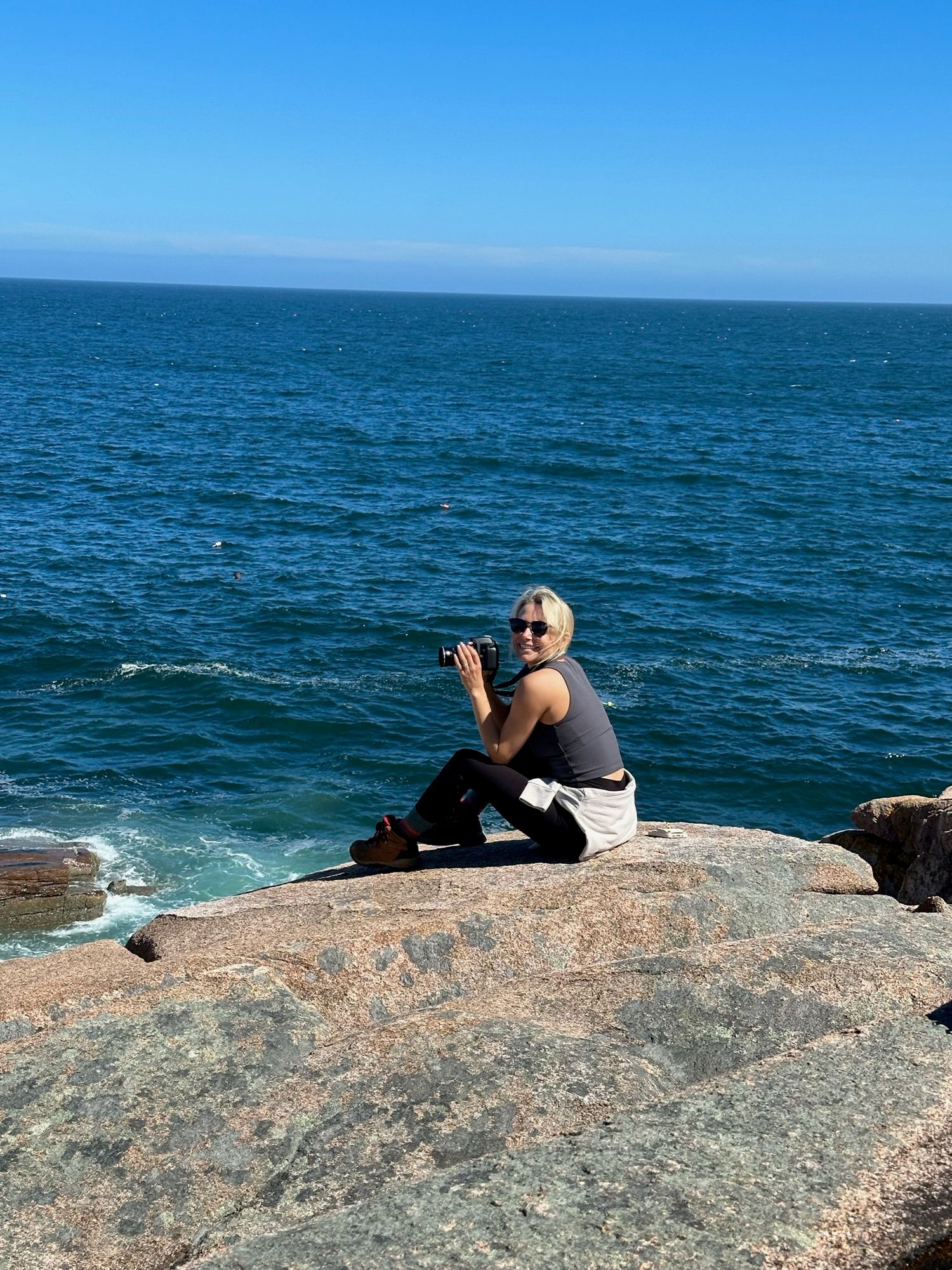 Photographer sitting on granite rocks by the sea on a sunny day with blue ocean backdrop.