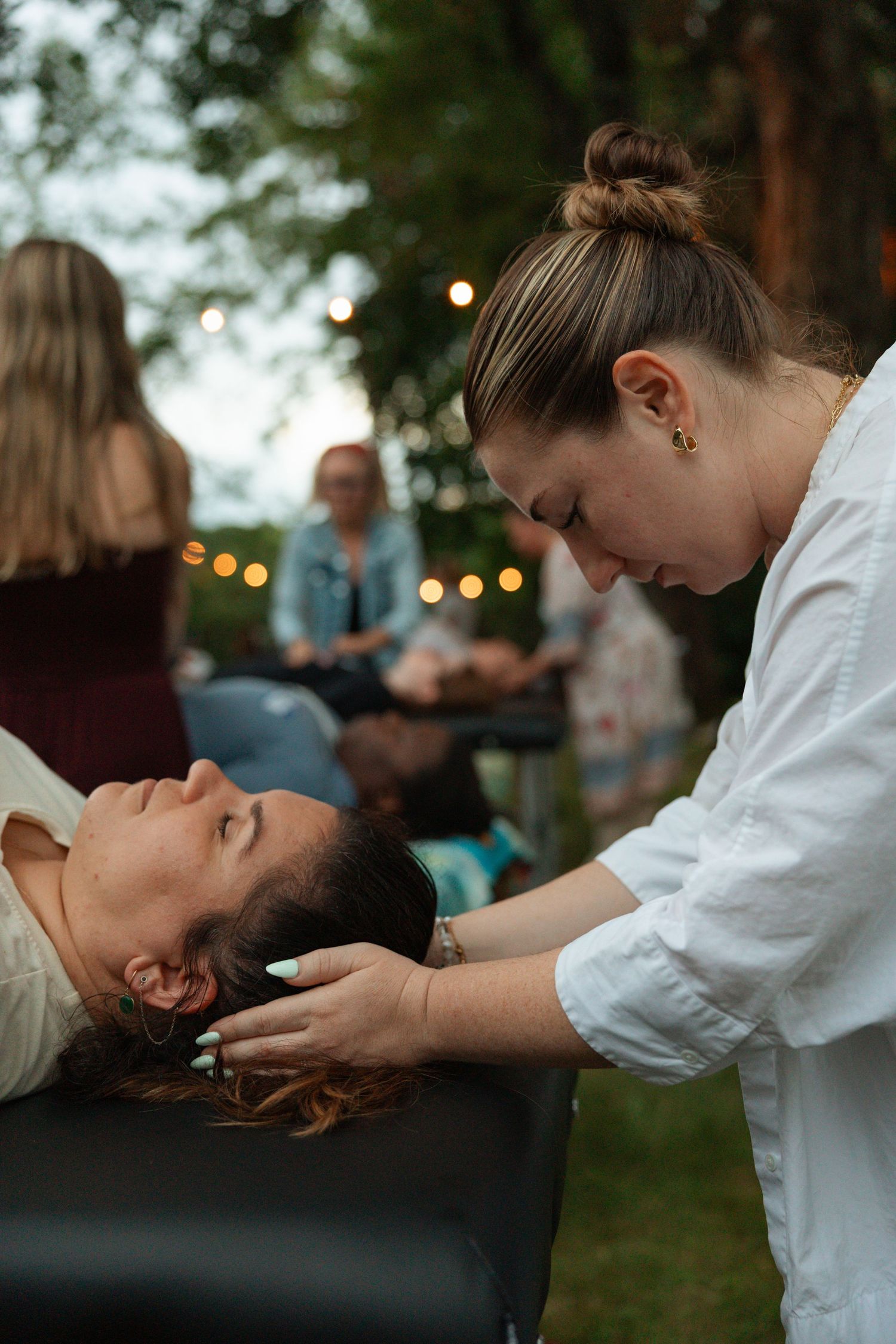 Woman receiving an outdoor head massage on a table at a relaxing evening wellness event with string lights.