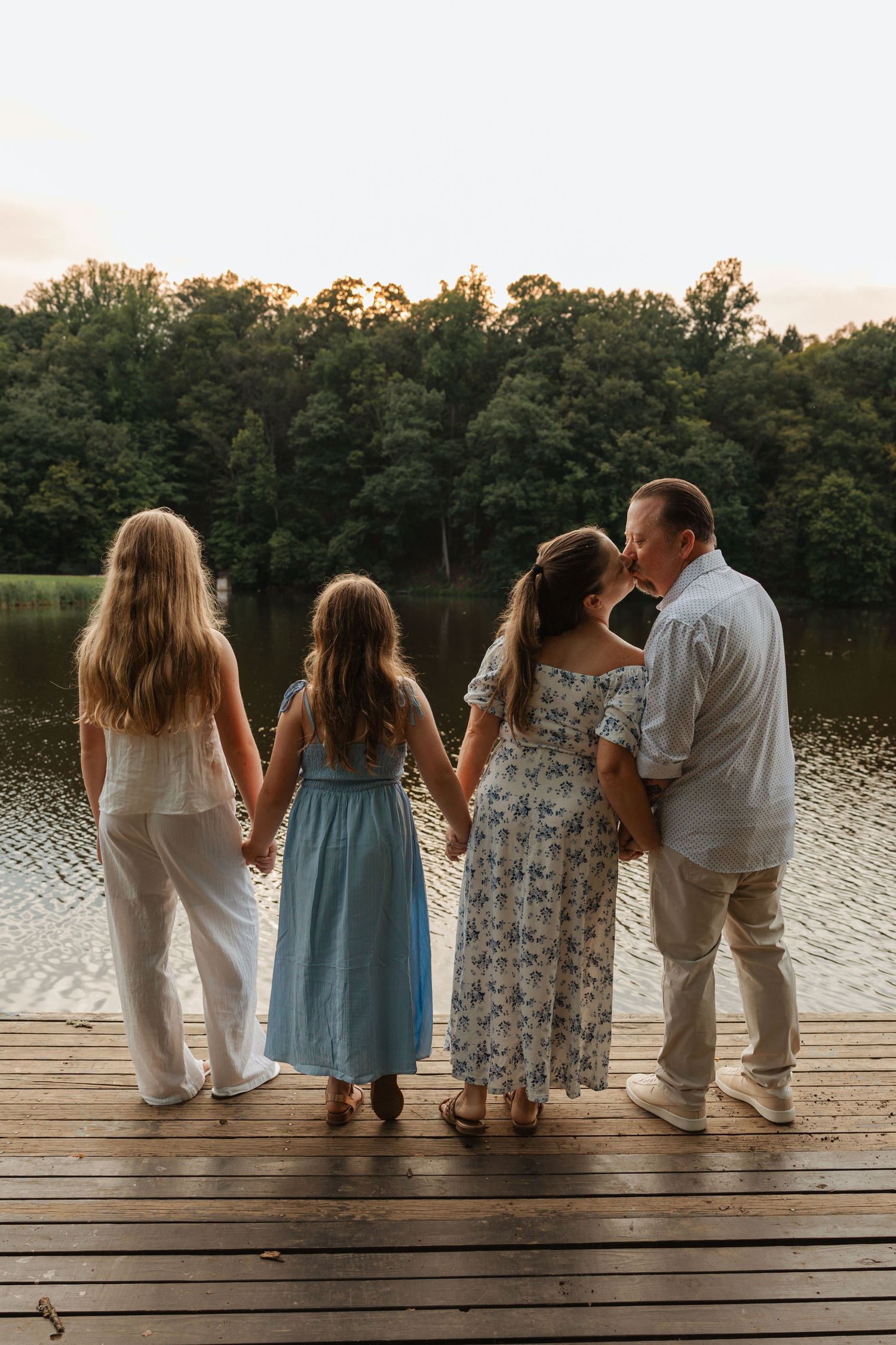 Family of four holding hands on a wooden dock at sunset, facing a serene lake surrounded by lush green trees.