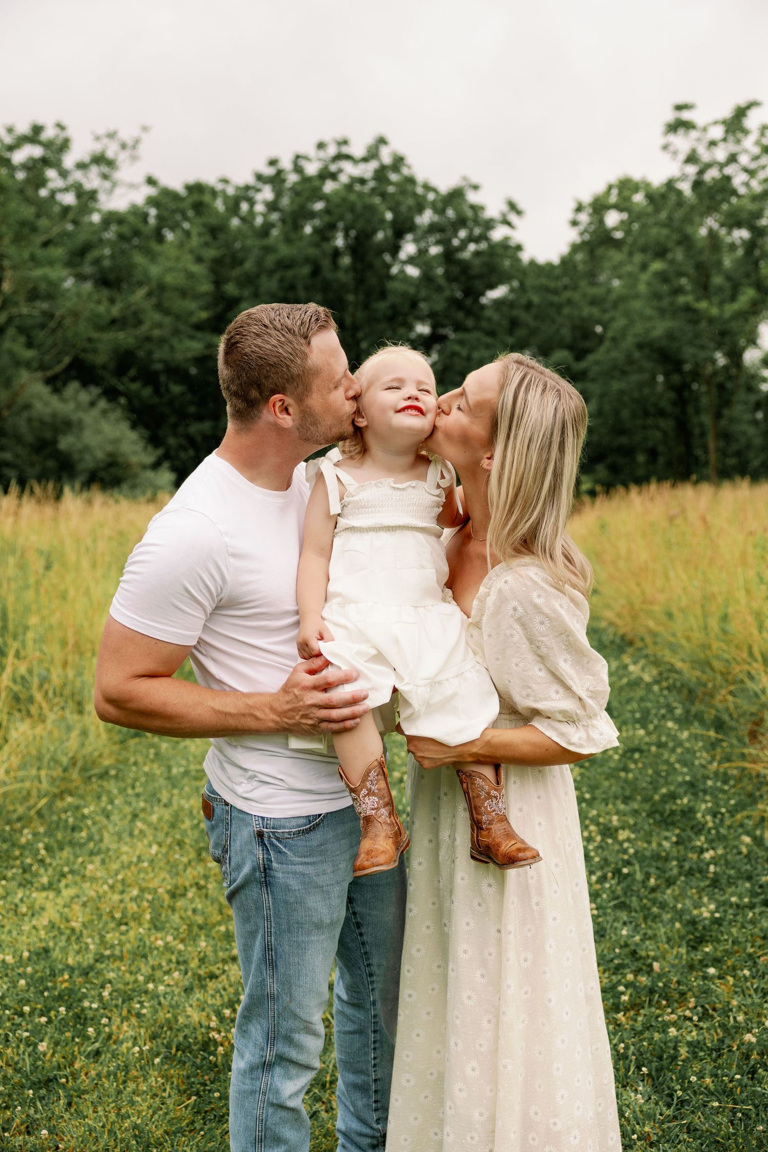 A couple kisses their smiling toddler girl in a white dress outdoors in a green field.