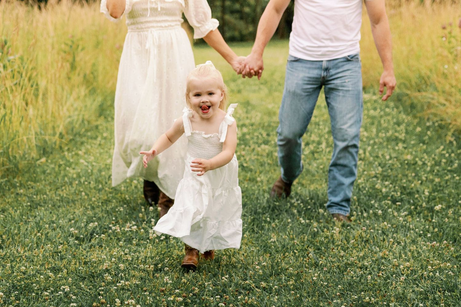 Joyful toddler girl in white dress laughs while walking with parents in a green field.