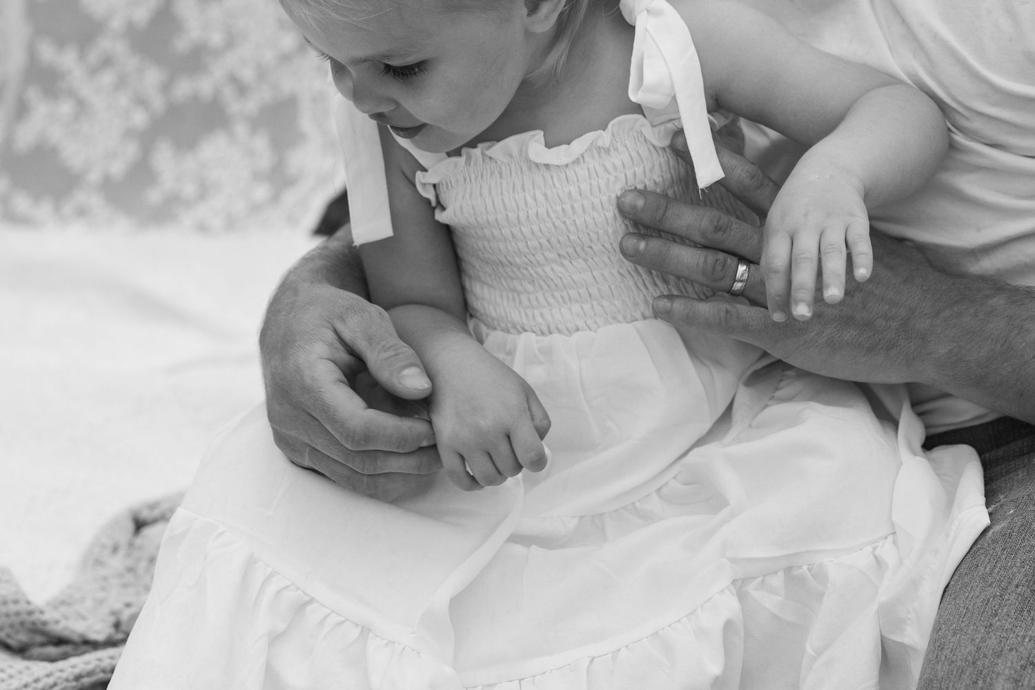 Close-up black and white photo of a baby in a white dress with an adults hands gently holding her.