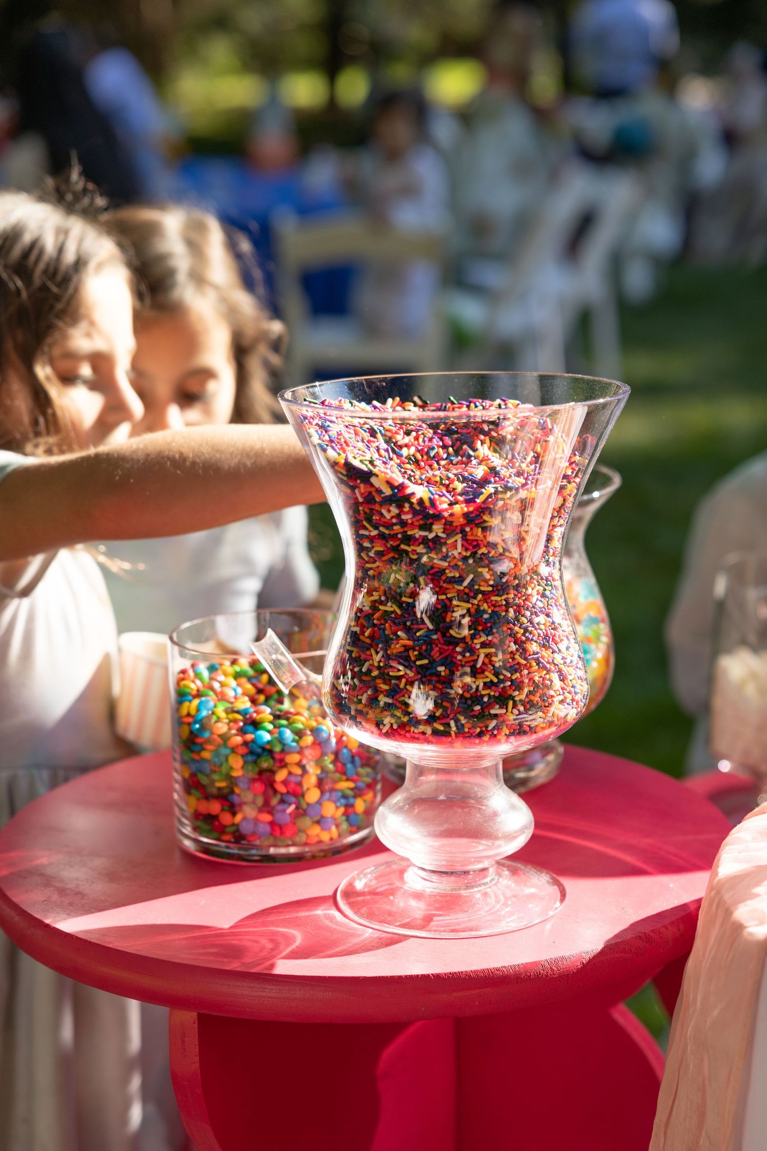 Children reaching into large glass vases filled with colorful rainbow sprinkles at an outdoor party.