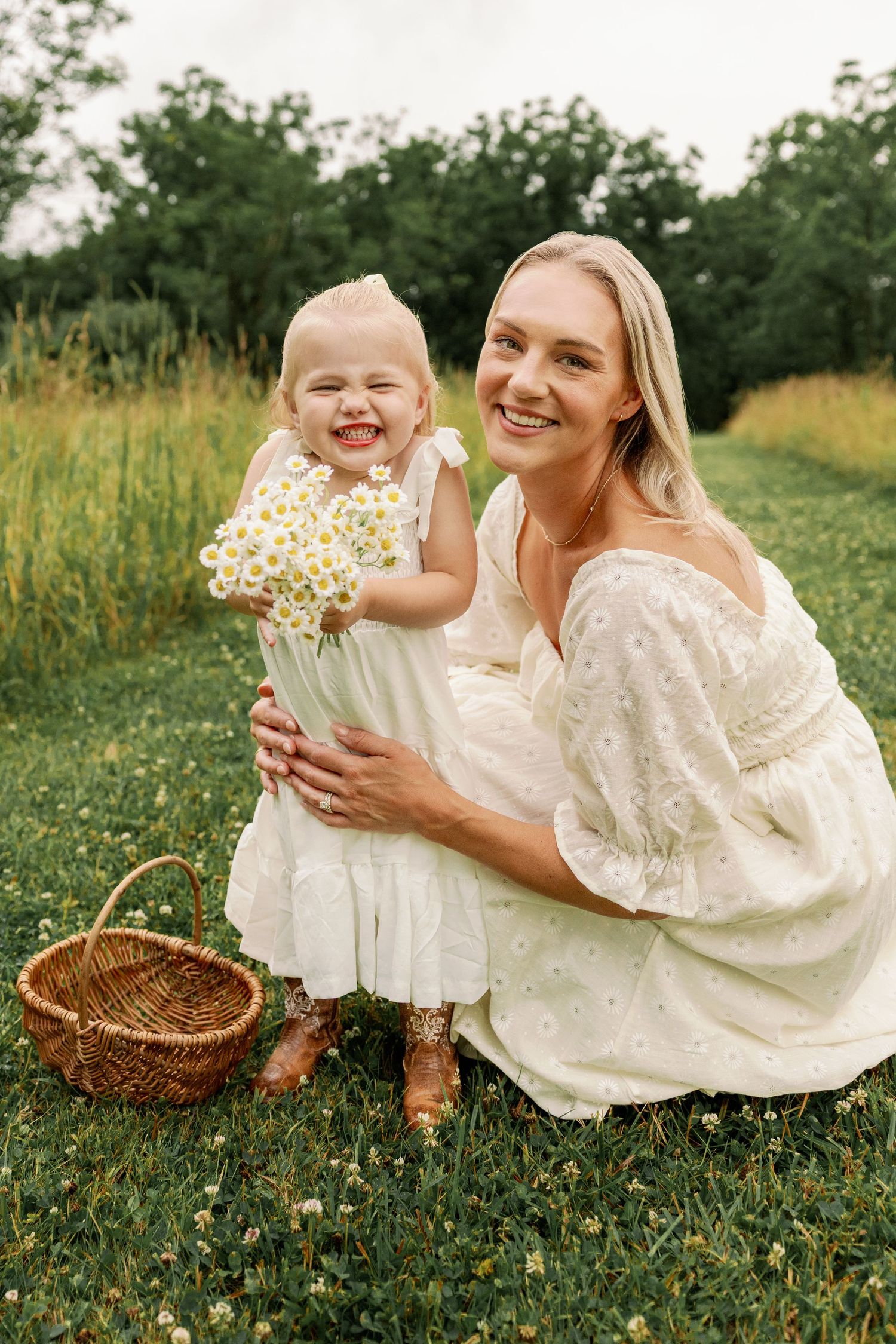 Smiling mother crouches beside laughing toddler daughter holding daisies with wicker basket in field.