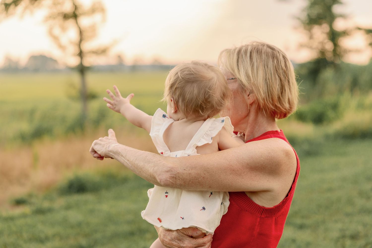 Blonde woman in red dress holding a baby outdoors in a golden sunset field setting.