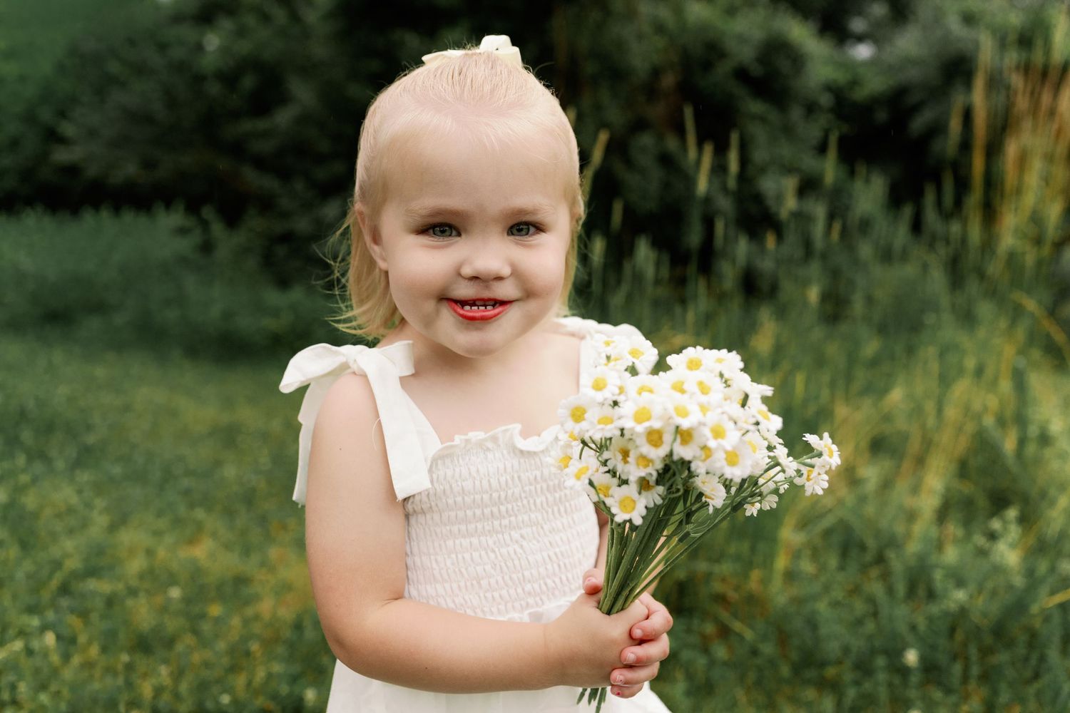 Adorable blonde toddler girl in white dress holds bouquet of white daisies in a green field.