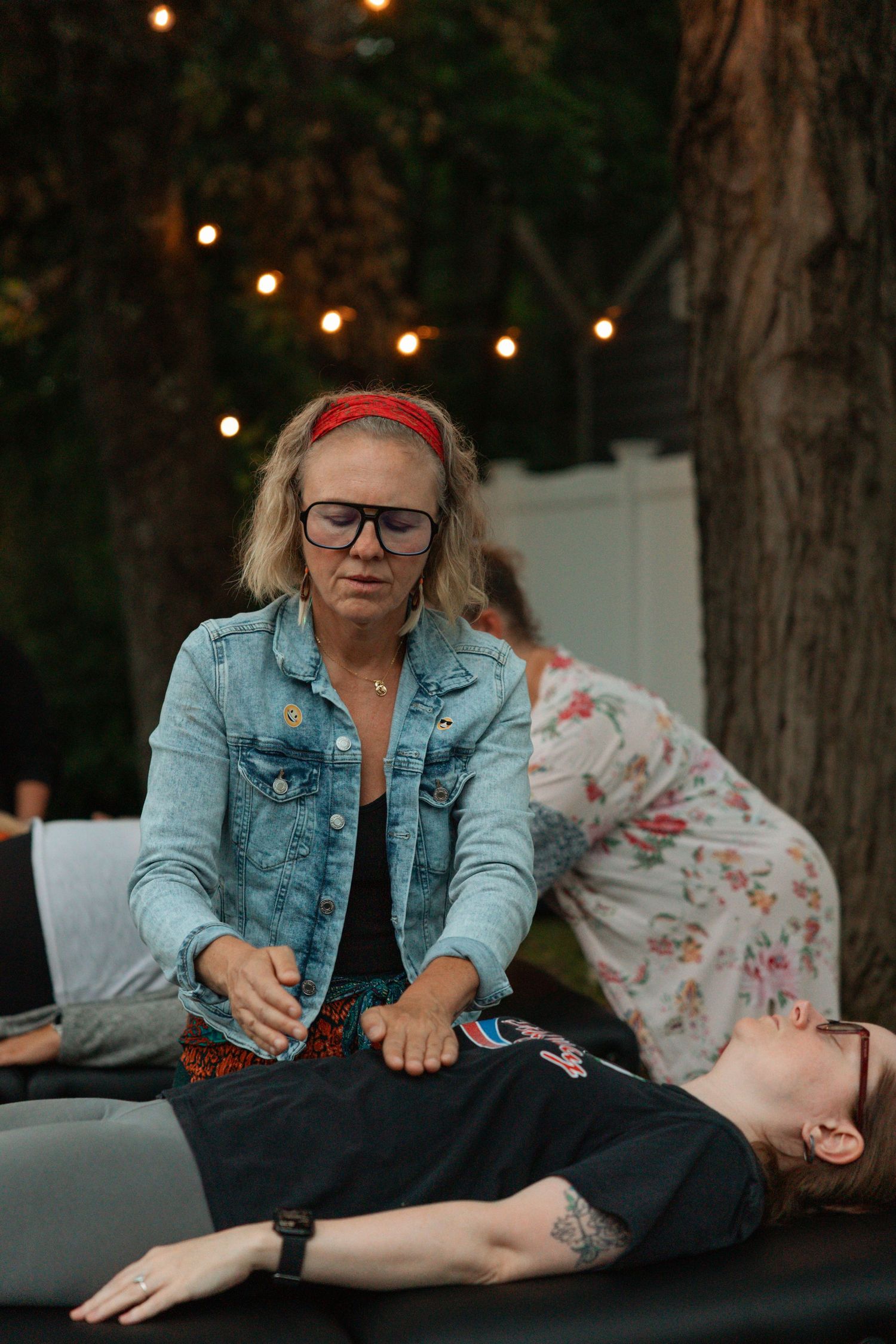 Woman in denim jacket giving outdoor massage therapy session at evening event with string lights.