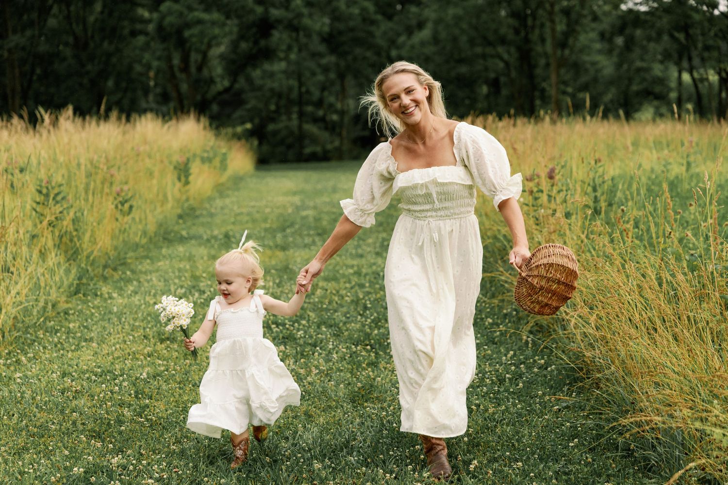 Happy mother in white dress walks hand-in-hand with toddler daughter through a scenic green meadow.