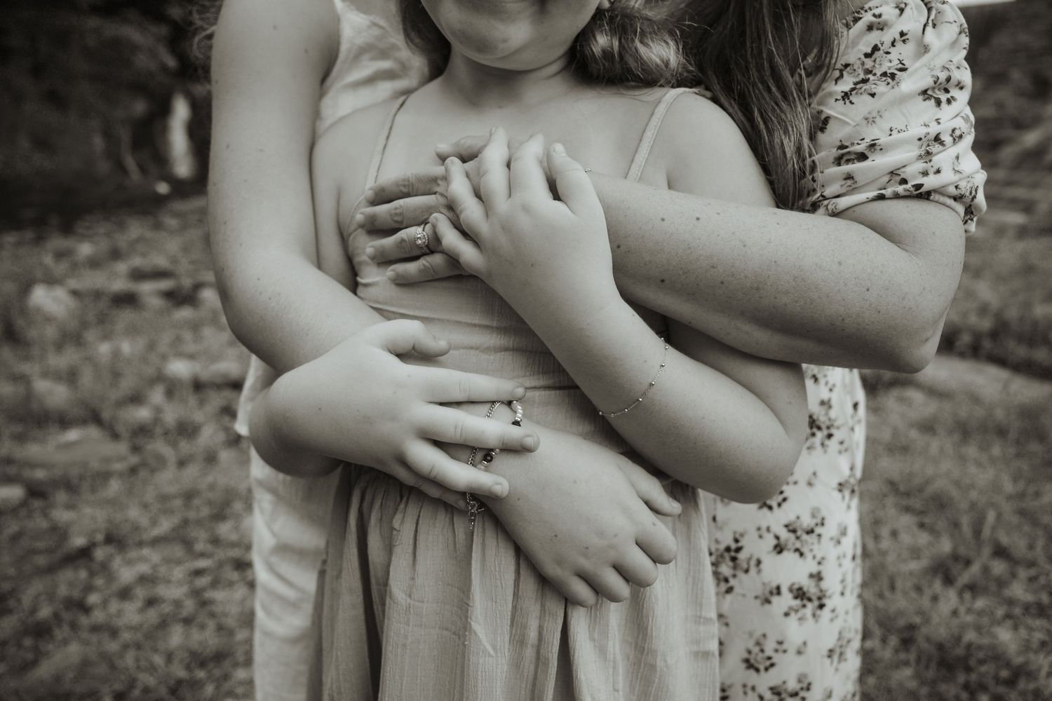 Black and white photo of two girls embracing, one wrapping arms around the other in a tender outdoor moment.