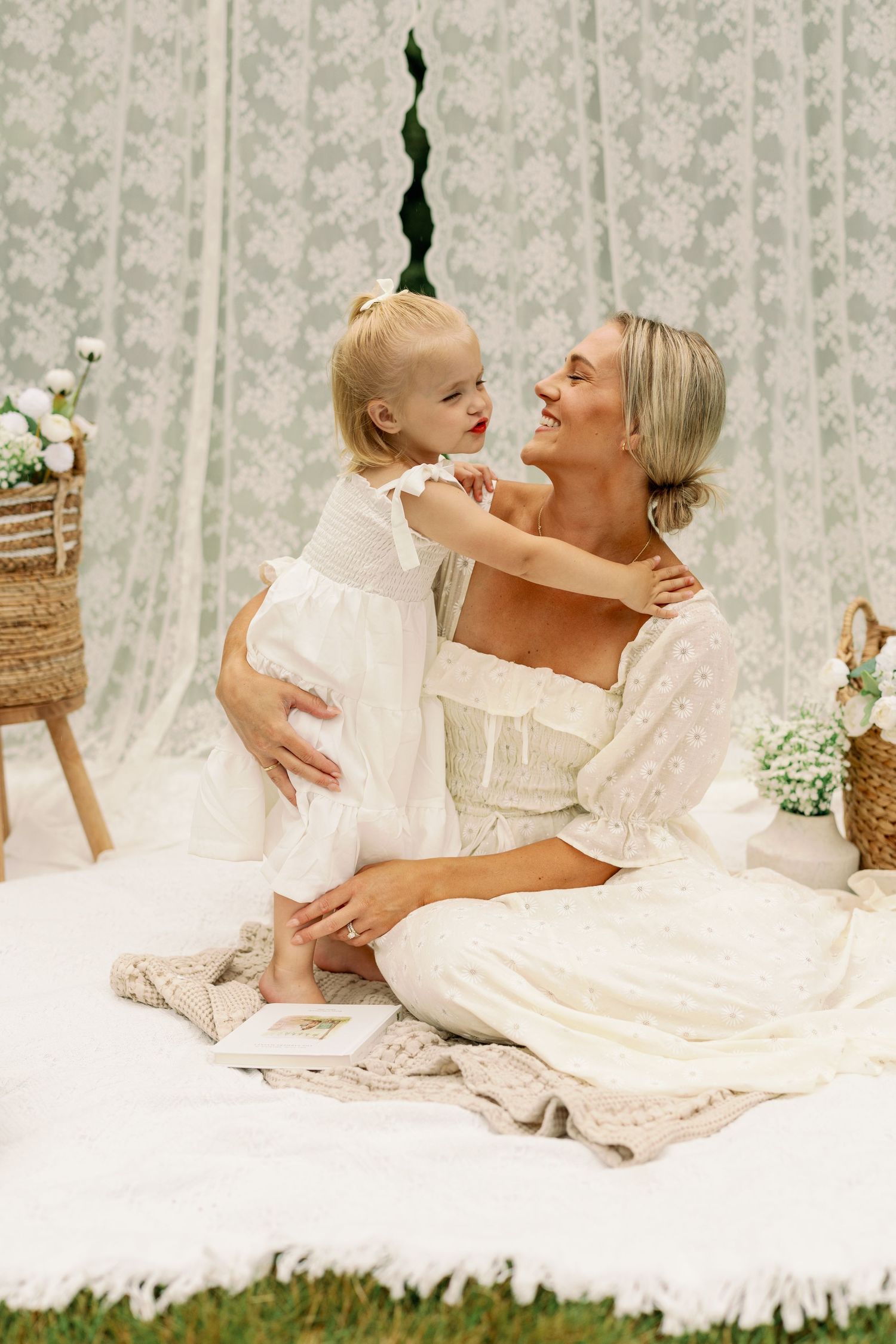 Smiling mother in white eyelet dress sitting with toddler daughter on blanket with floral decor.