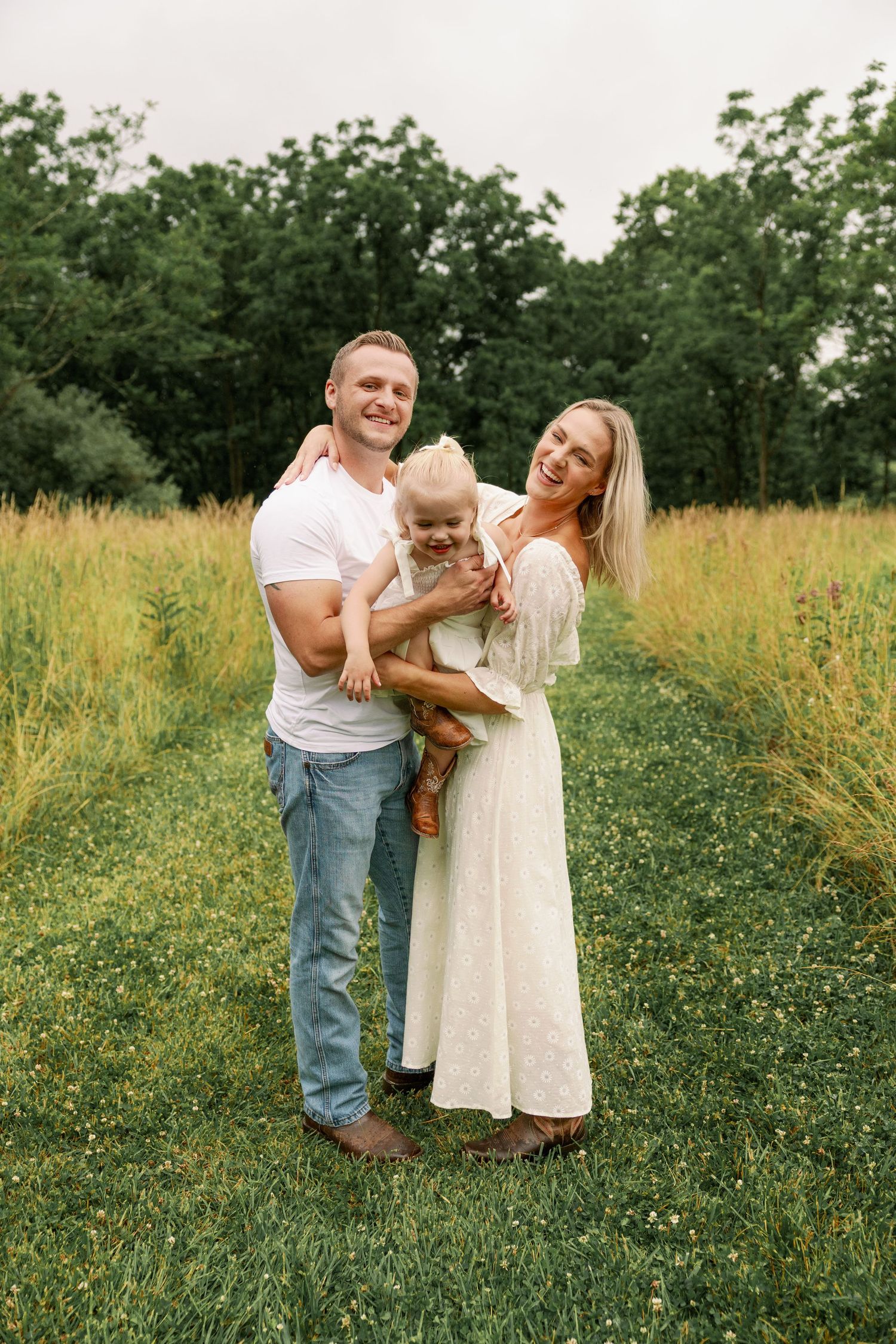 Smiling family of three with baby enjoys a sunny day in a green grassy field.
