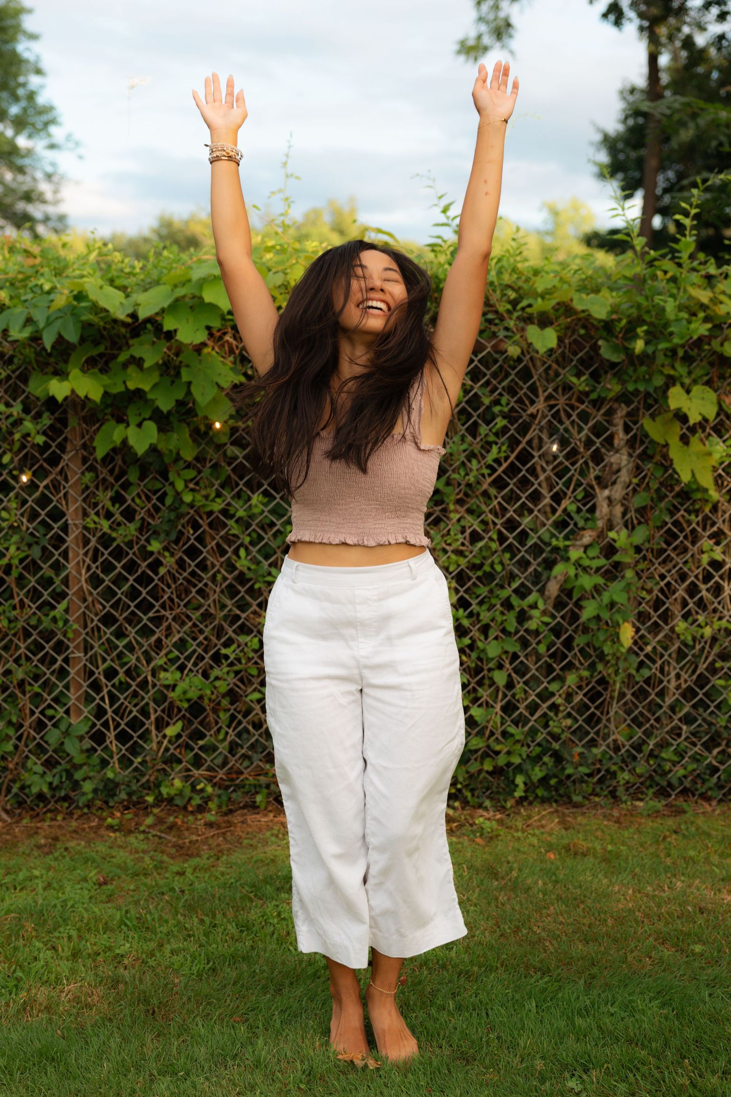 Joyful young woman with arms raised outdoors, wearing a beige crop top and white wide-leg pants in a lush garden.