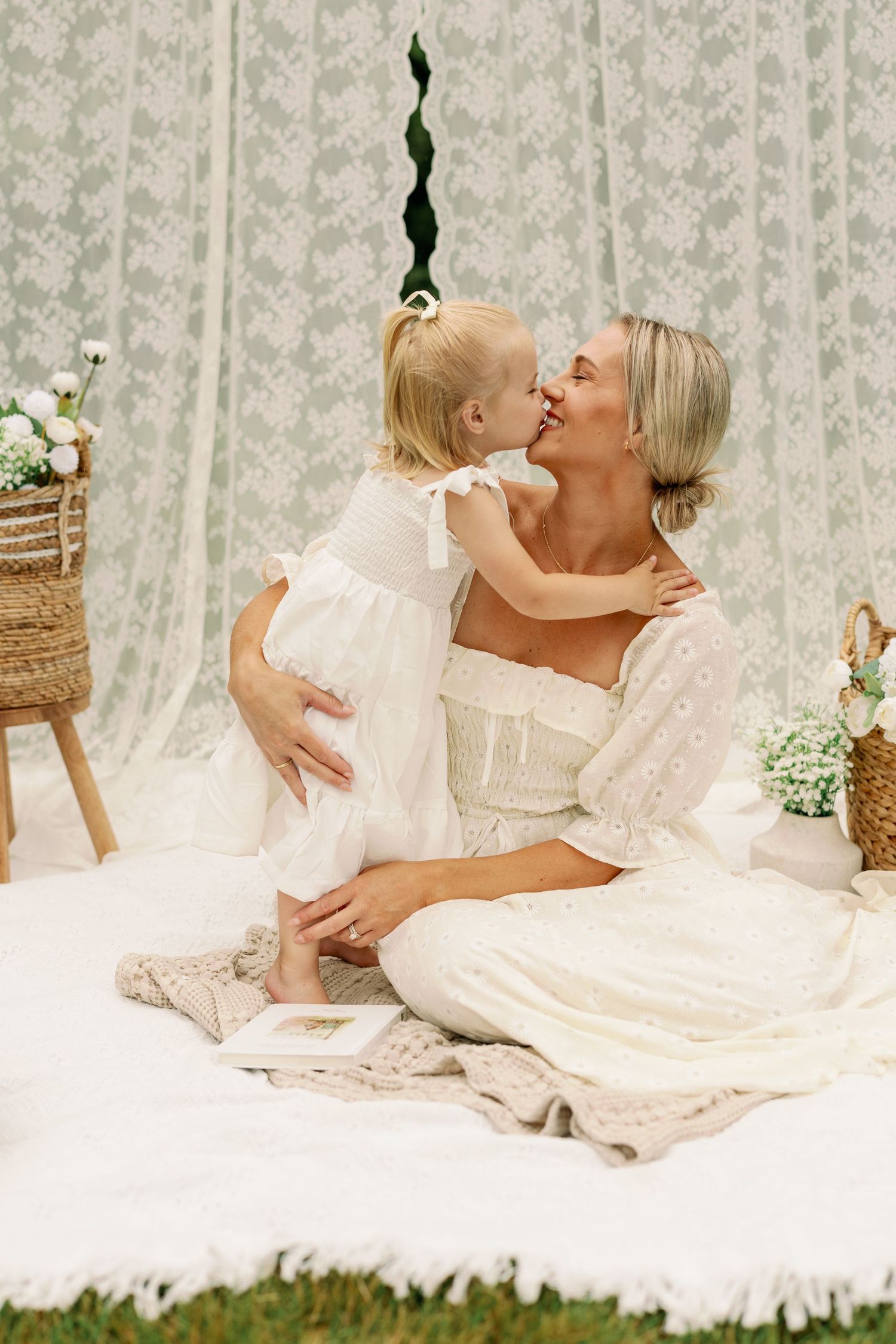 Mother and toddler daughter sharing a sweet kiss on white blanket with lace backdrop outdoors.